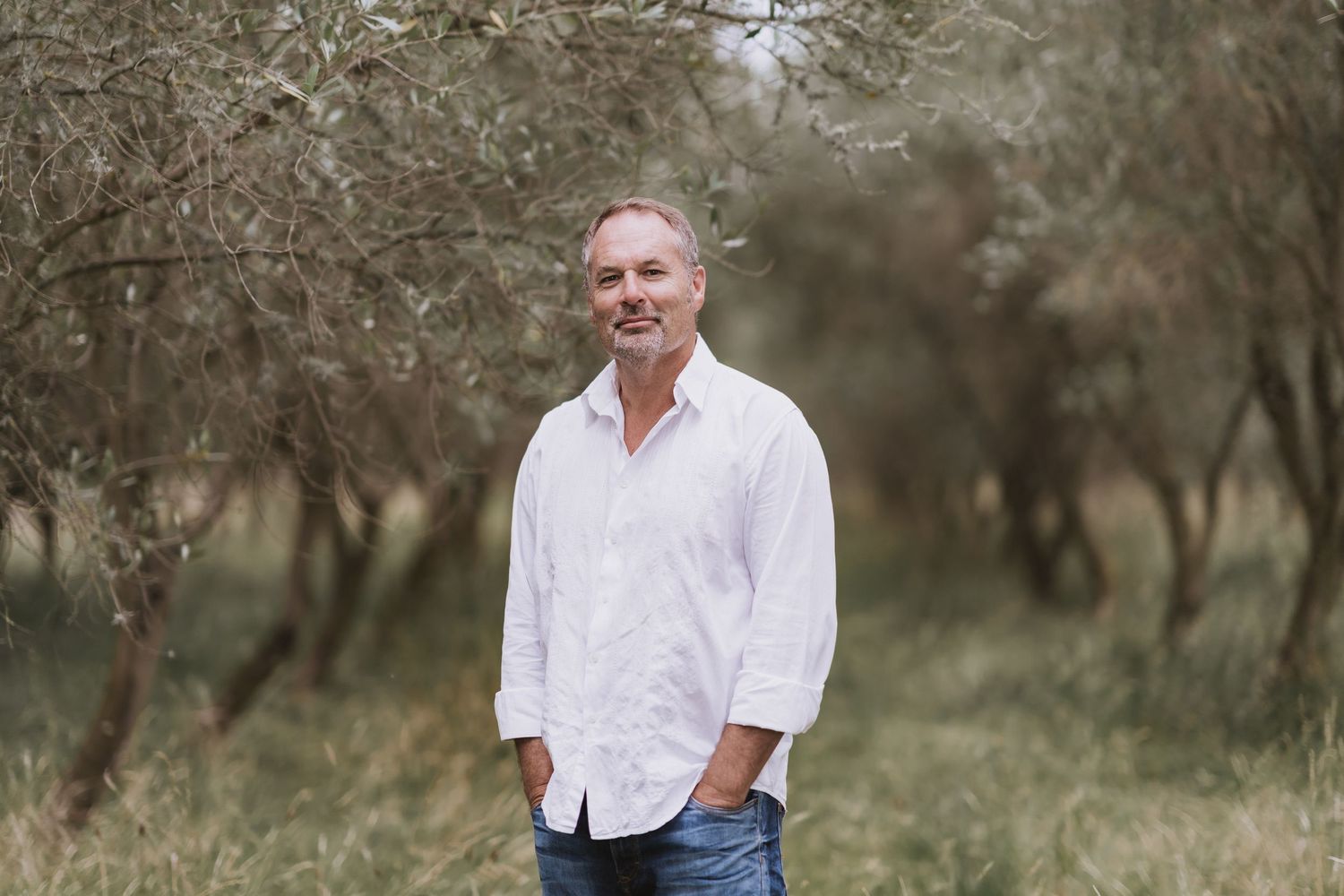Person in white shirt posing naturally in an olive grove setting with natural desaturated earth tones.