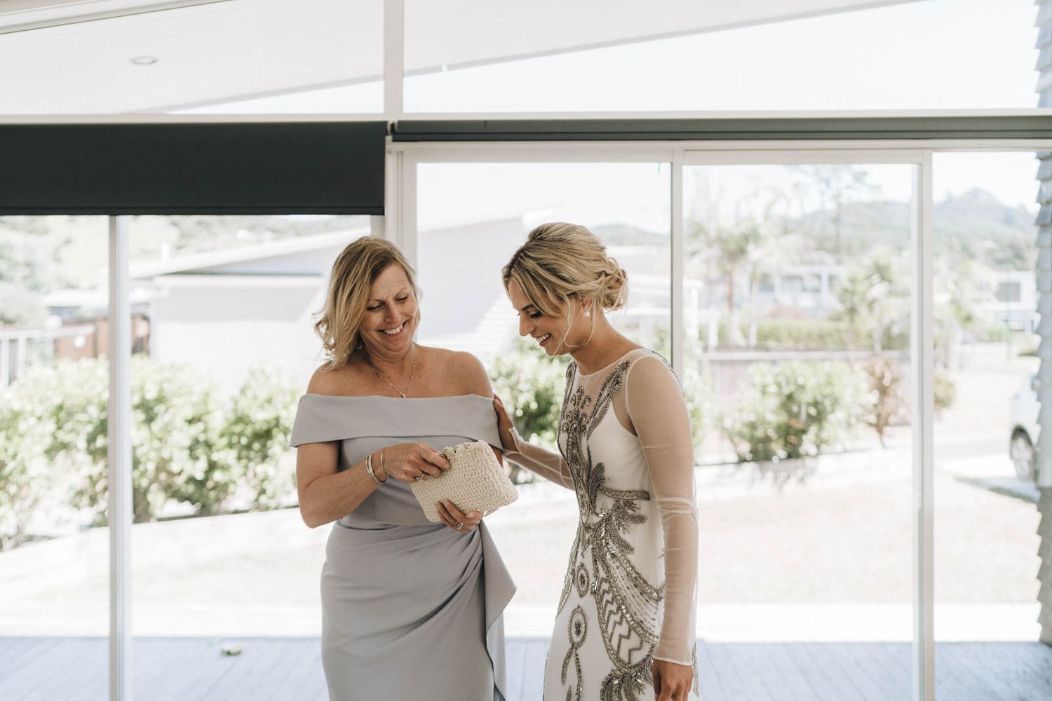 Two wedding attendees share a close conversation in elegant grey and floral dresses by large windows.