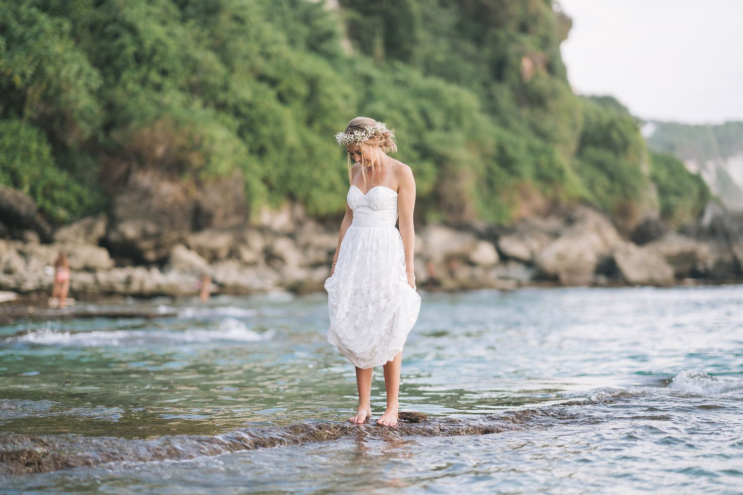 Bride in white dress standing in shallow ocean water with cliffs in background.
