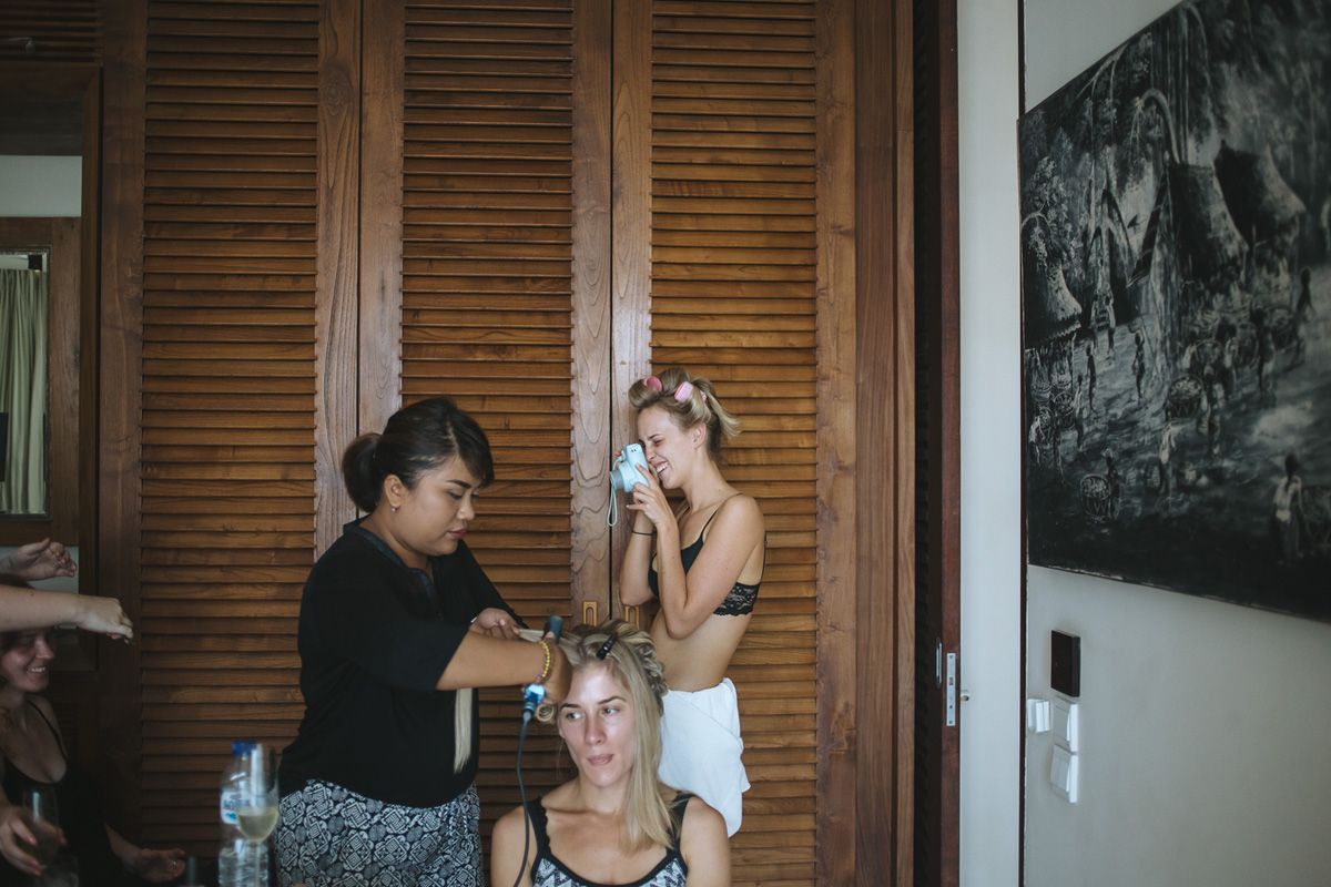 Two people helping a third person with makeup preparations in a room with wooden shutters and a landscape photo on the wall.