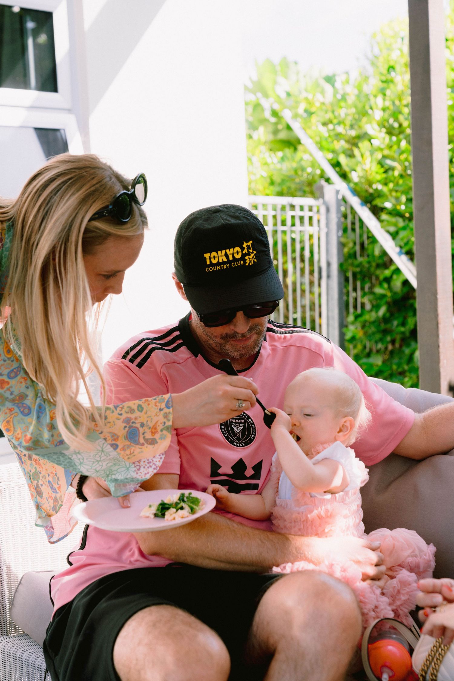 Family members share a sweet moment on porch, feeding and spending time with a baby in a pink outfit.