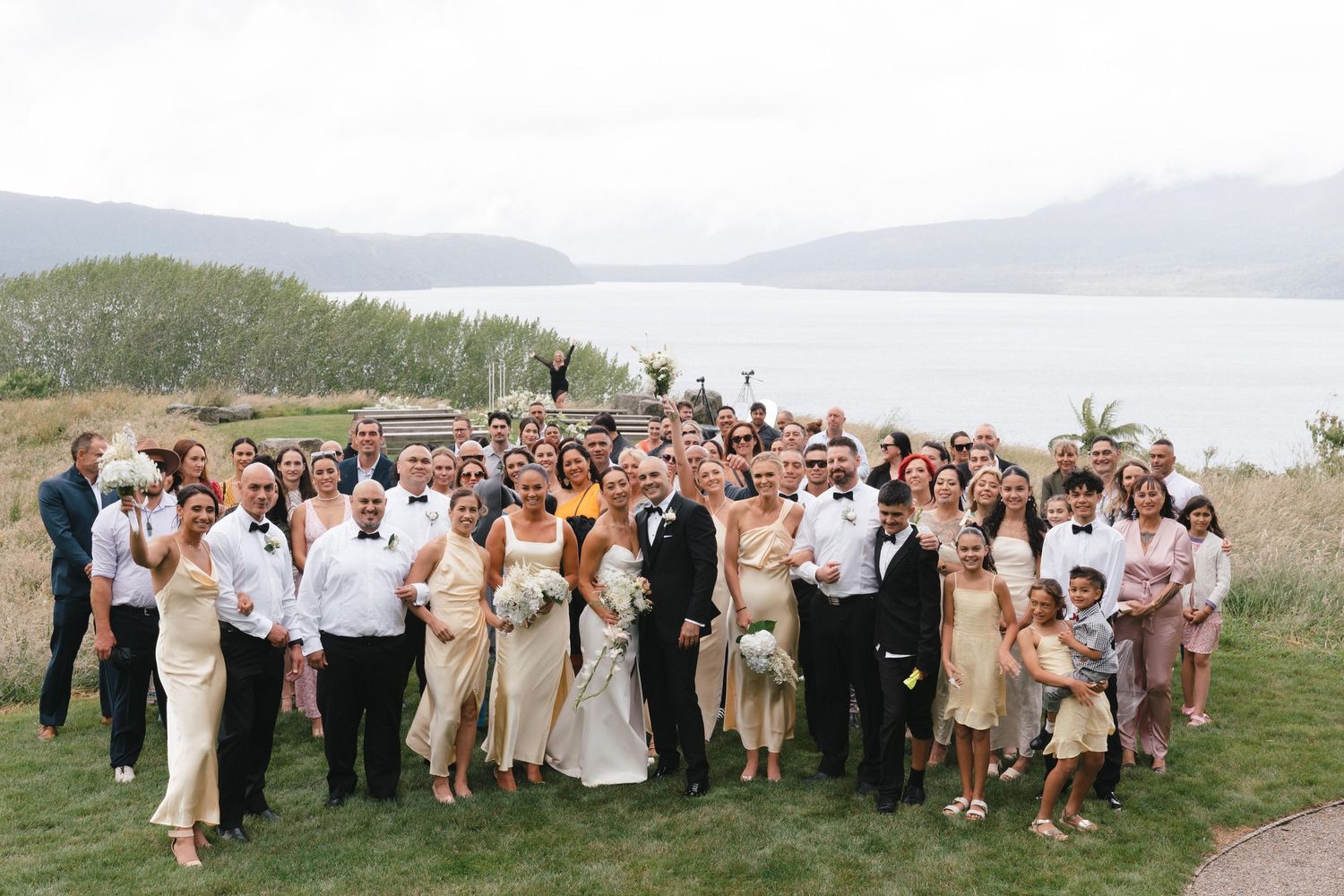 Large wedding party poses for a panoramic group photo on a hillside with water views.