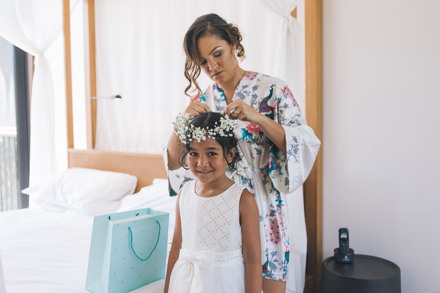 A floral robe-clad figure styles someone's hair in a bright white room with a blue shopping bag.