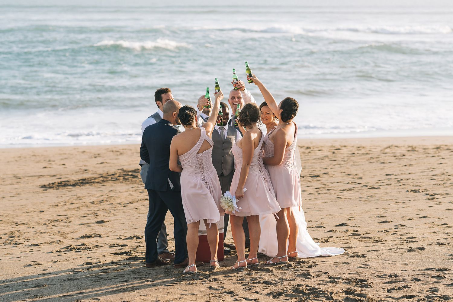 Group celebrating with raised bottles on a beach at sunset while ocean waves crash in the background.