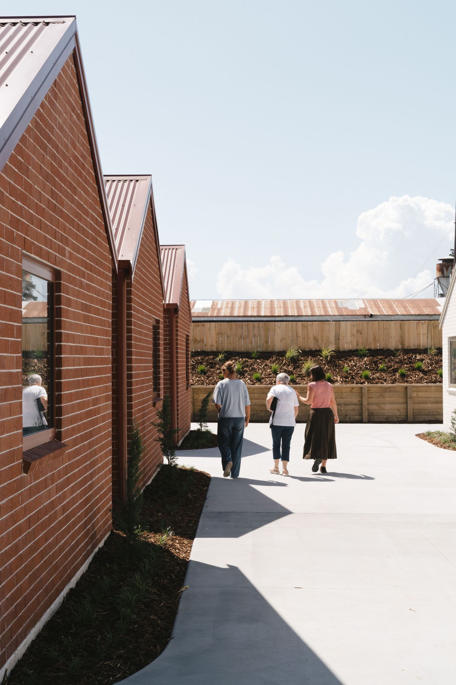 People walking along a concrete path between modern brick buildings with metal roofing on a sunny day.