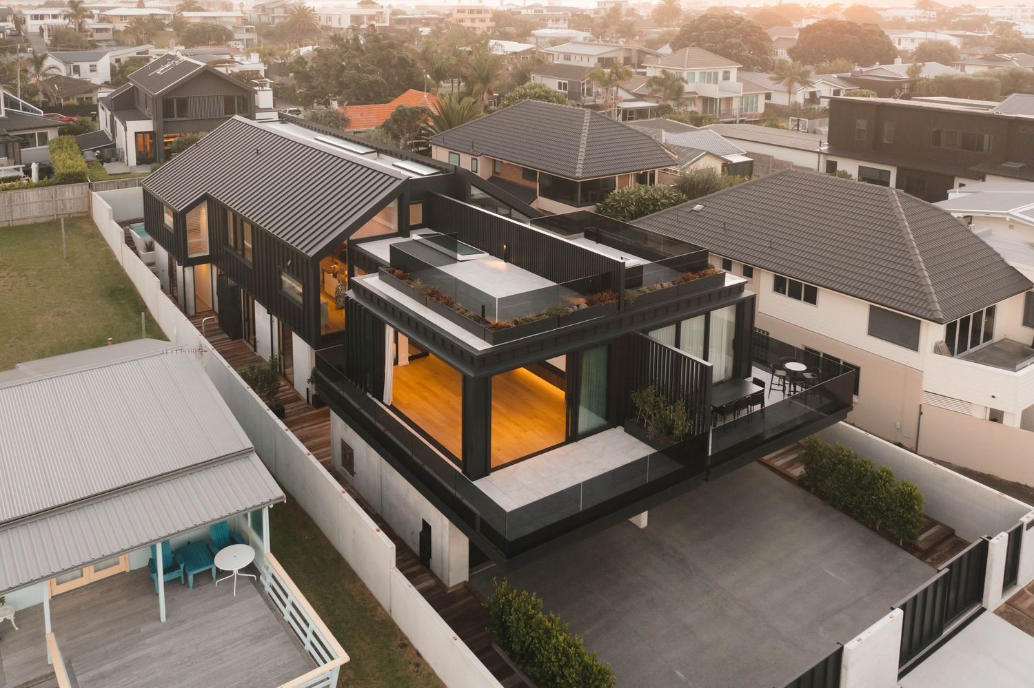 Modern black house with glass walls and orange interior lighting glows at dusk in suburban neighborhood.