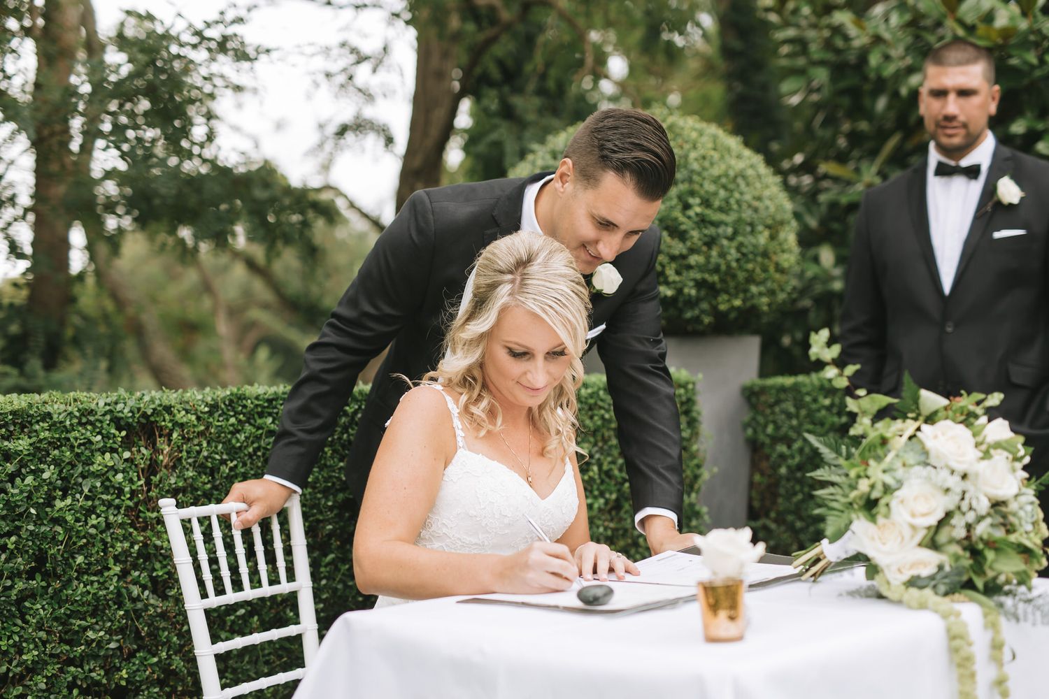 Wedding party celebrates at outdoor reception tables decorated with lush greenery and white flowers in garden setting.