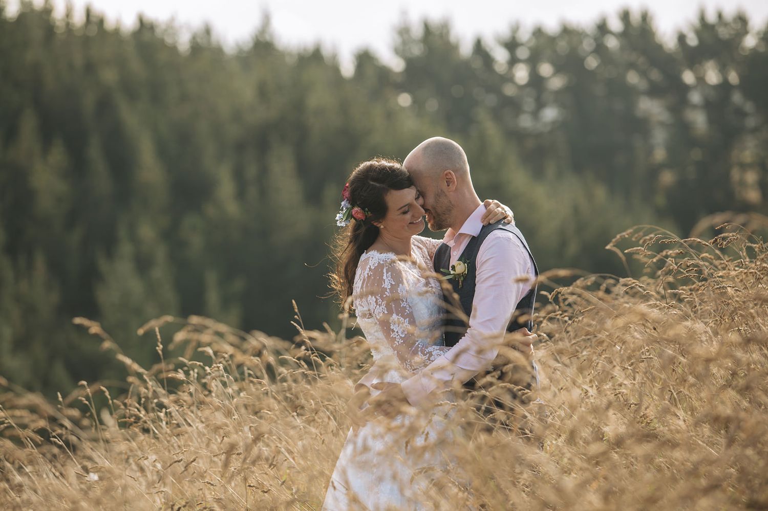 Romantic couple embraces in golden sunlit wheat field with evergreen forest backdrop during sunset wedding photoshoot.