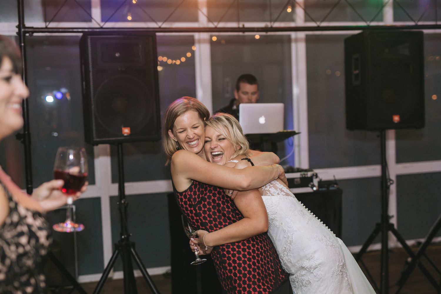 Dance floor scene at a wedding reception with people dancing and embracing in front of DJ equipment and string lights.