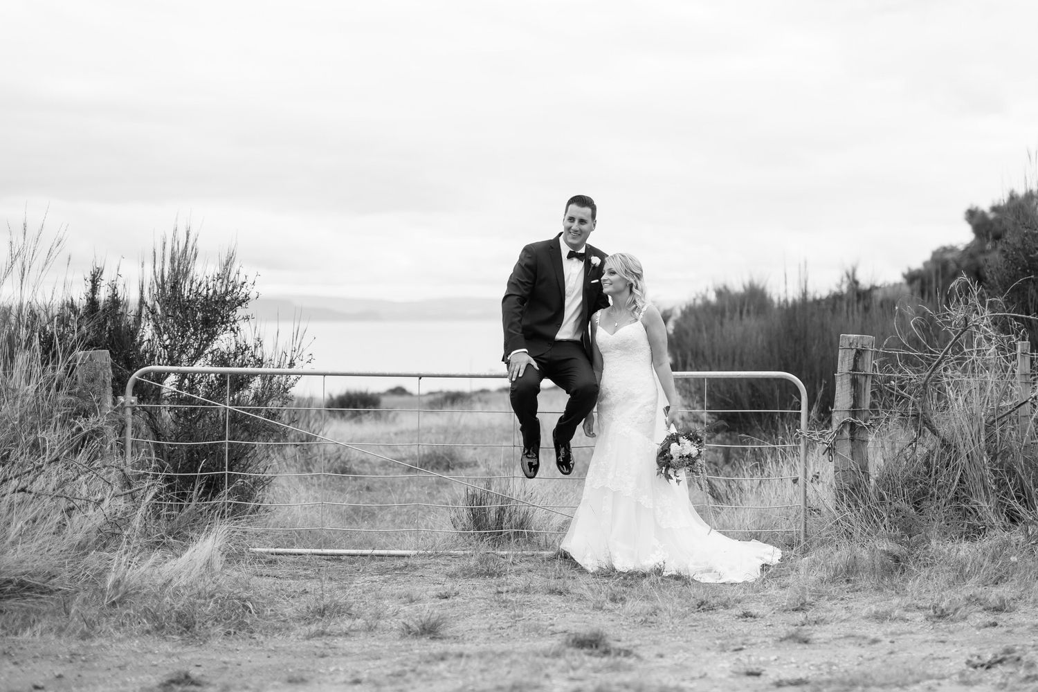 A couple in wedding attire stands by a metal gate on a rustic path overlooking a lake in black and white.