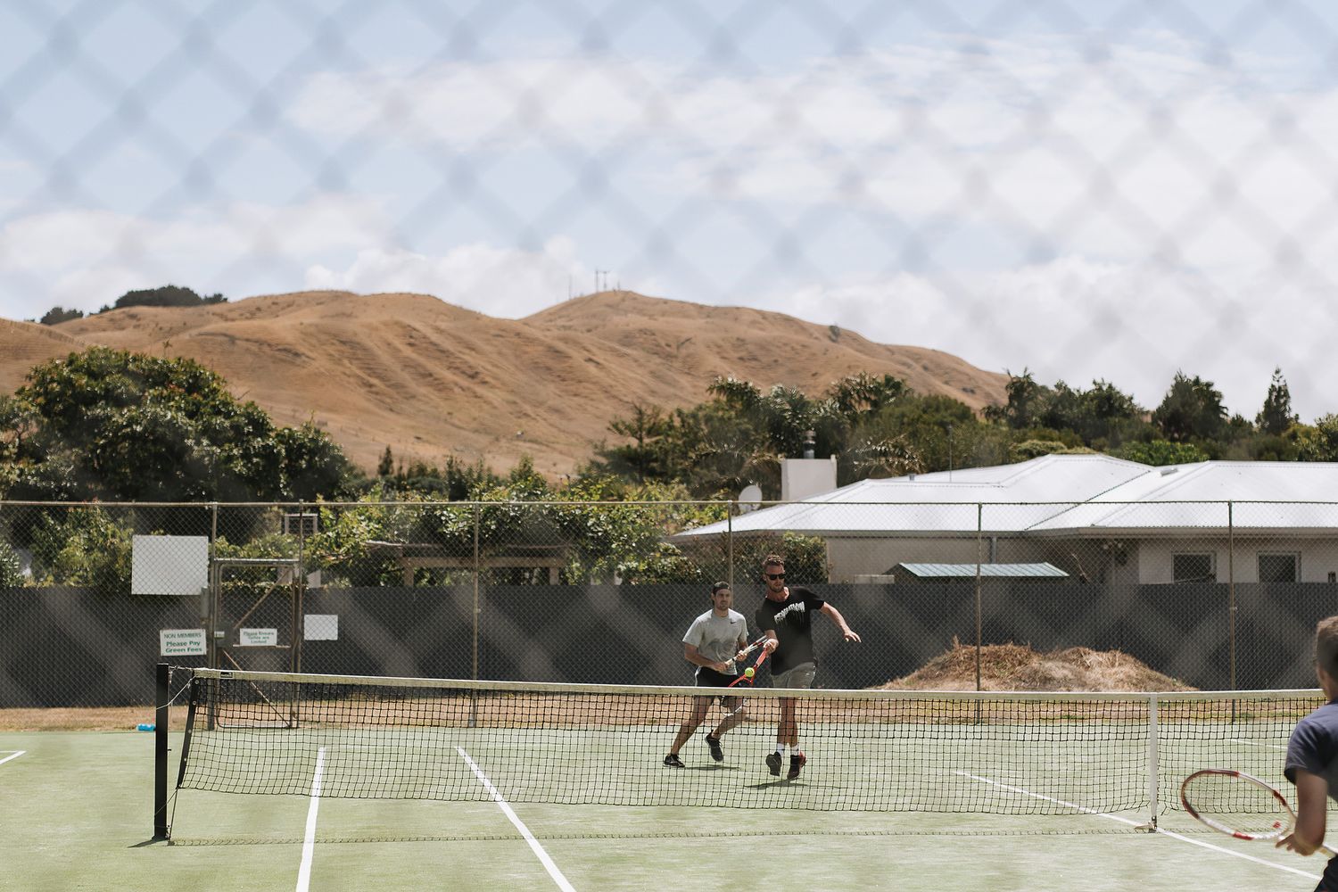 Tennis players warm up on clay courts surrounded by rolling brown hills and mountains in a scenic countryside setting.