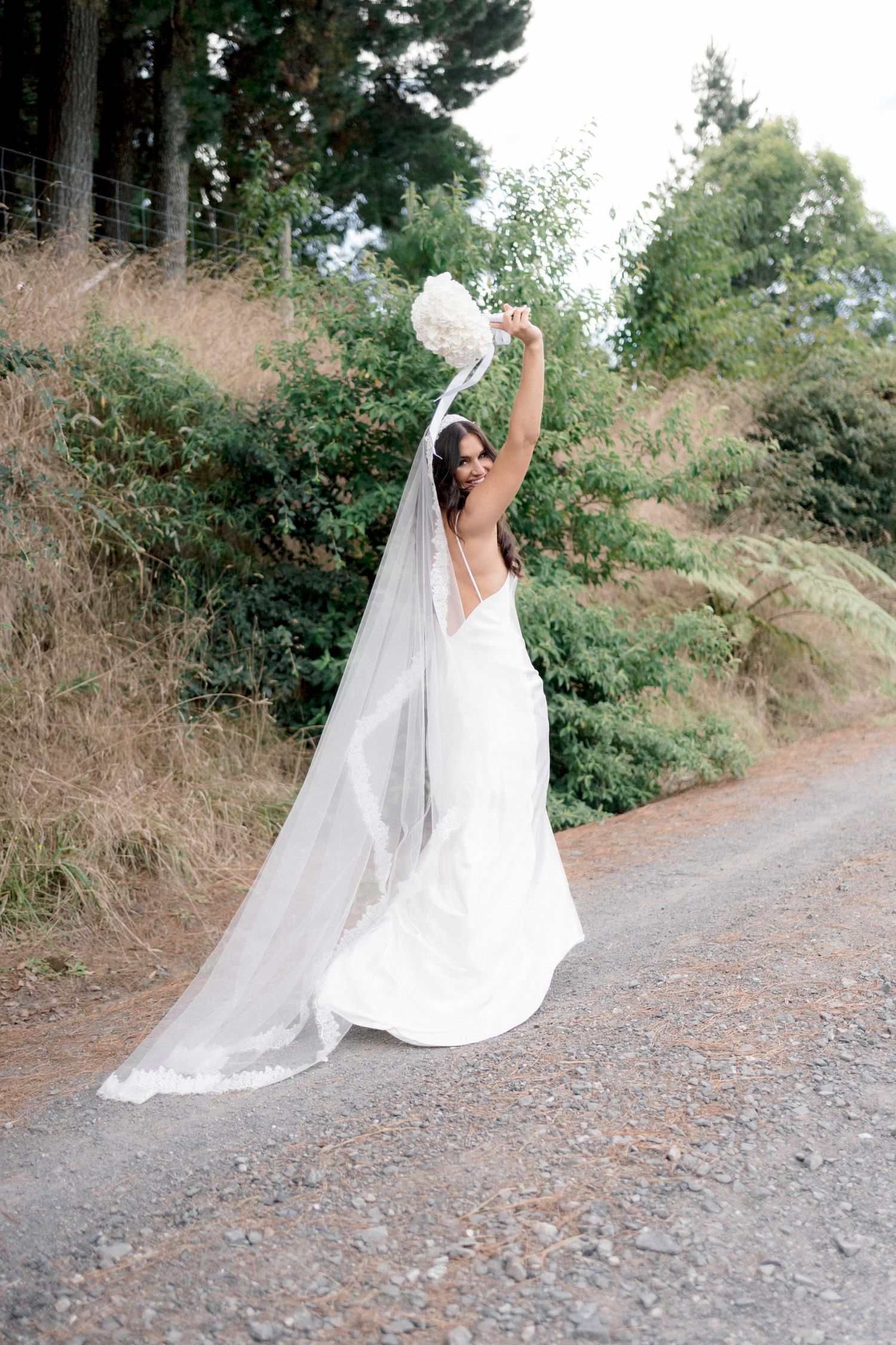 A bride in a flowing white dress and veil twirls joyfully on a dirt path surrounded by greenery.