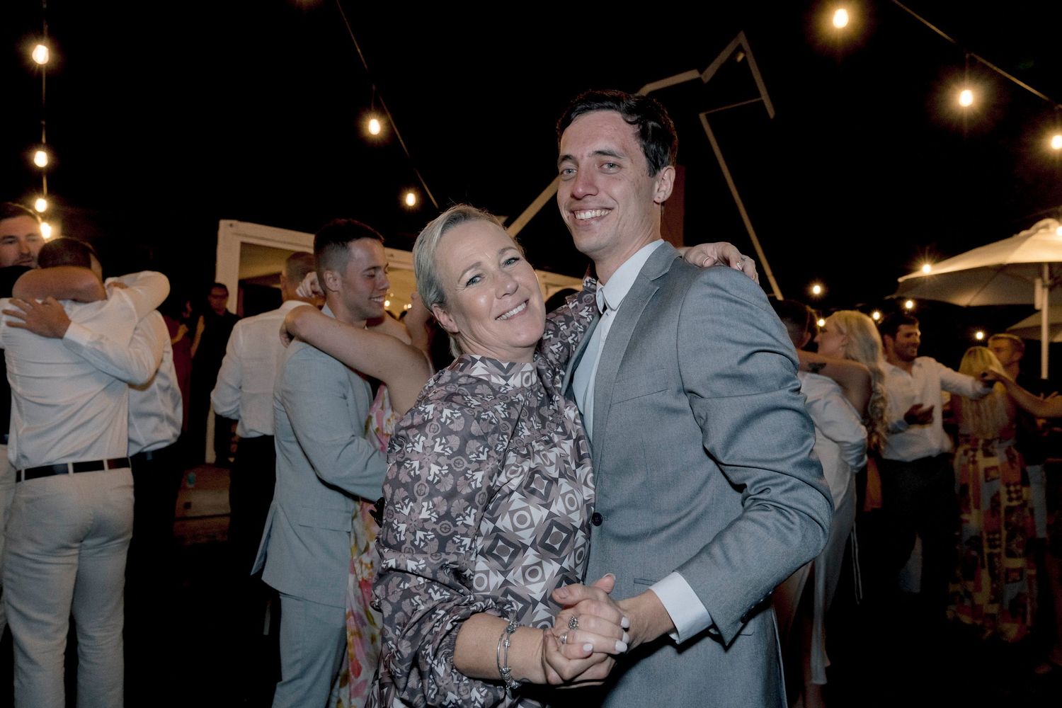 Wedding guests dance and celebrate under string lights at an outdoor evening reception.