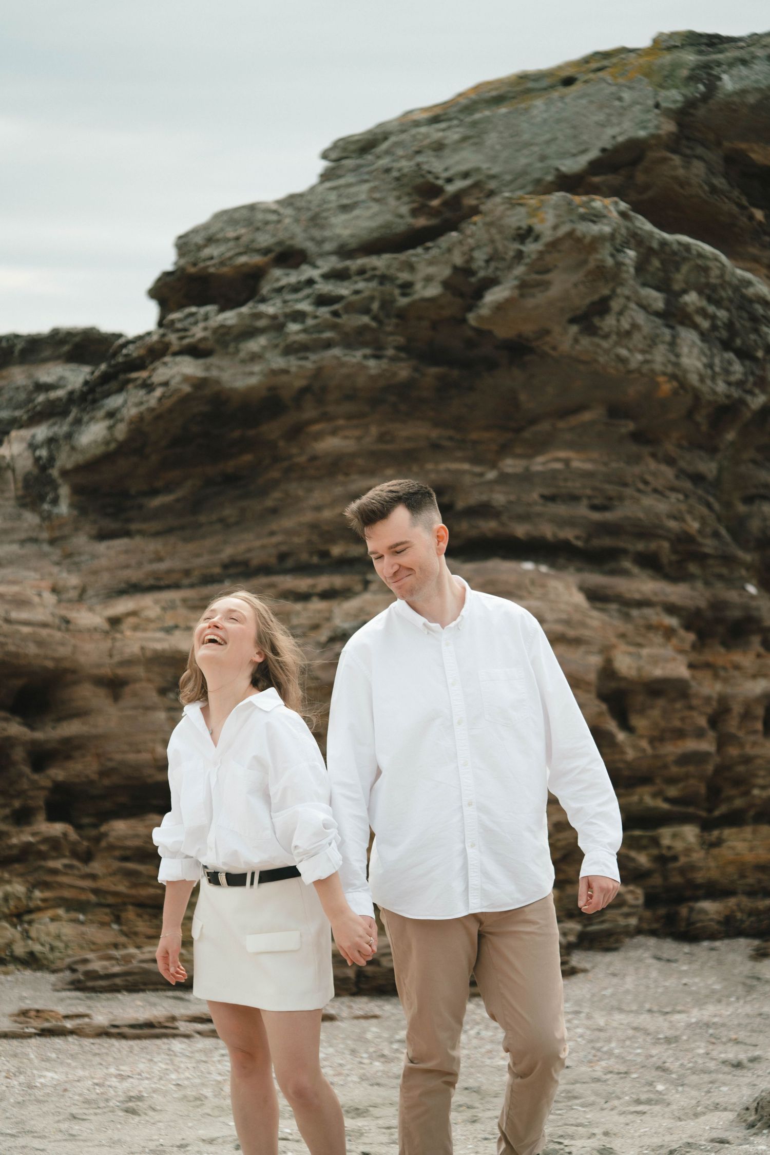 Couple in matching white attire walking hand in hand along a scenic rocky beach.