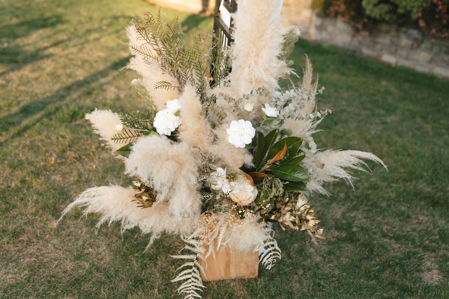 A rustic boho floral arrangement with pampas grass, white flowers and dried elements in a wooden box on grass.