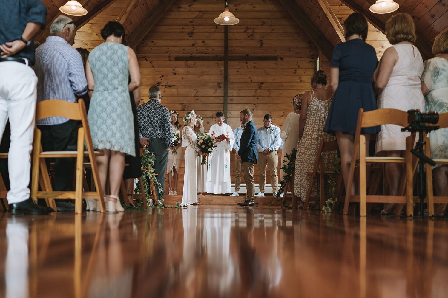 Wedding ceremony in a rustic wooden chapel with guests standing and seated on wooden chairs.