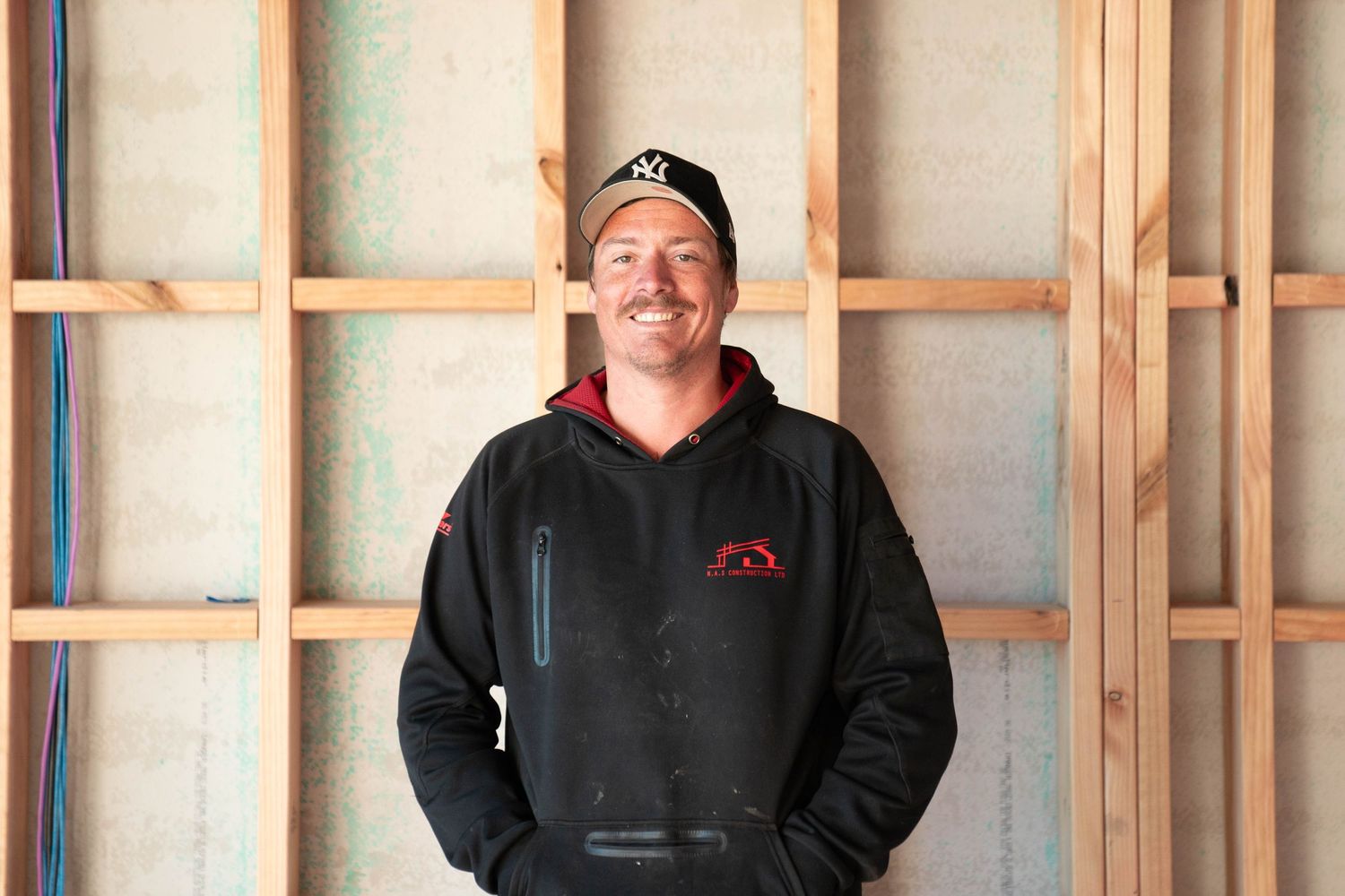 Construction worker in black hoodie smiling while standing in front of wooden framing in unfinished room.