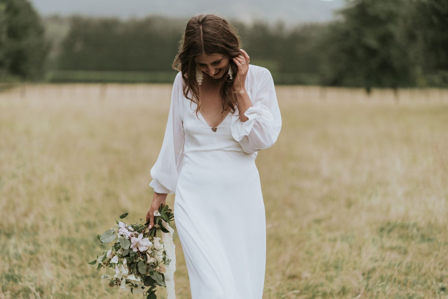 Person in a flowing white wedding dress stands in a golden field holding a wildflower bouquet on an overcast day.