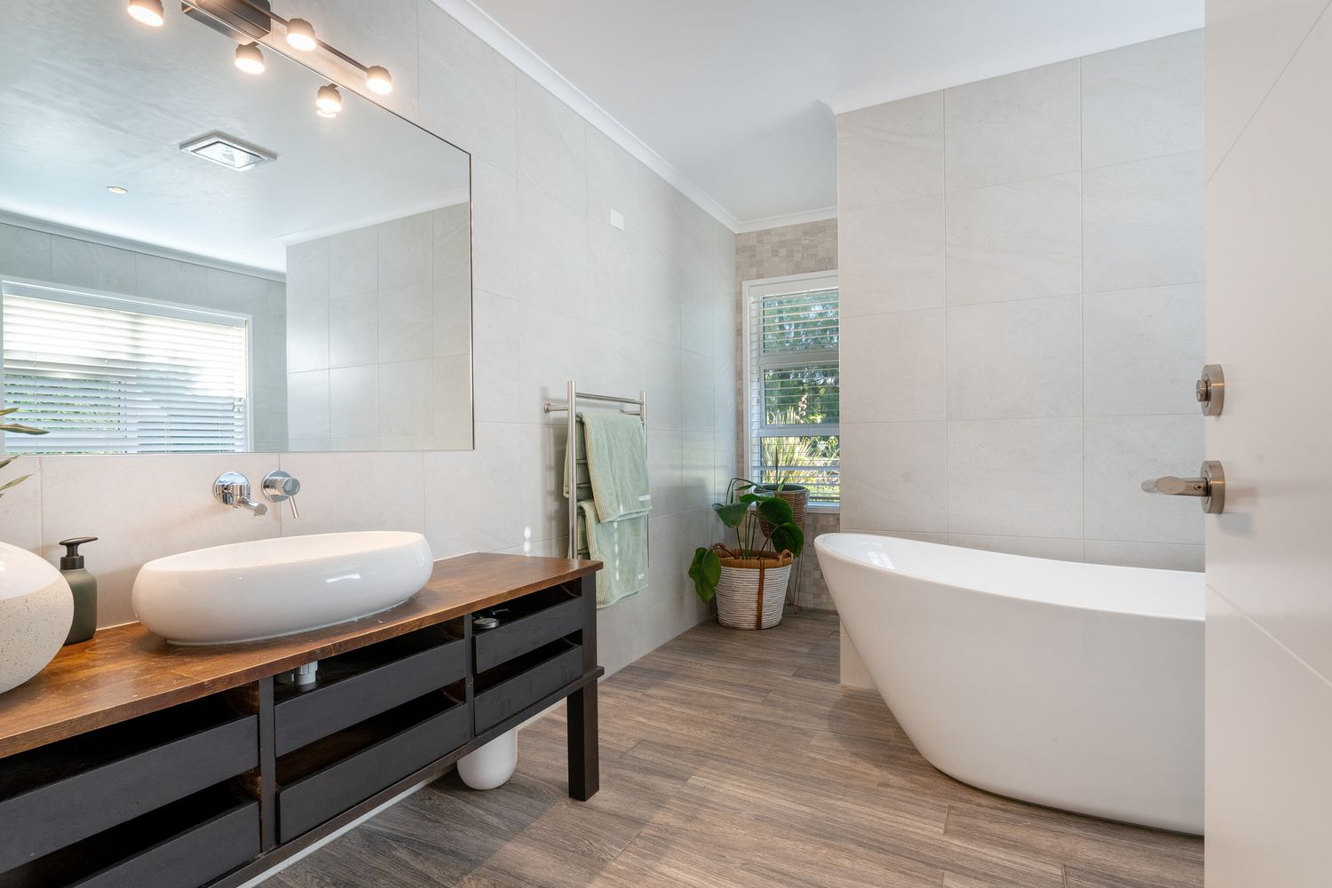 Modern bathroom featuring a freestanding tub, dual vessel sinks on wooden vanity, and wood-look flooring.