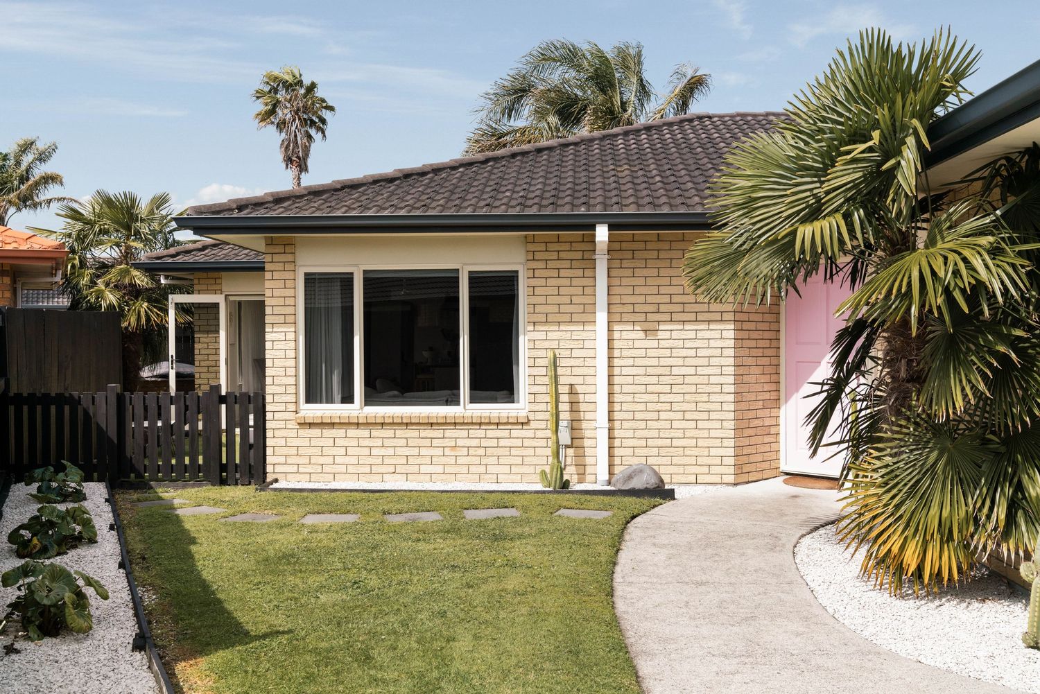 Modern single story brick home with palm trees and green lawn featuring curved concrete pathway and dark tiled roof.