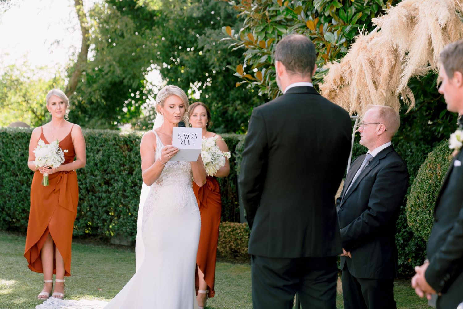 Wedding party poses outdoors in garden wearing formal attire including white dresses and suits.