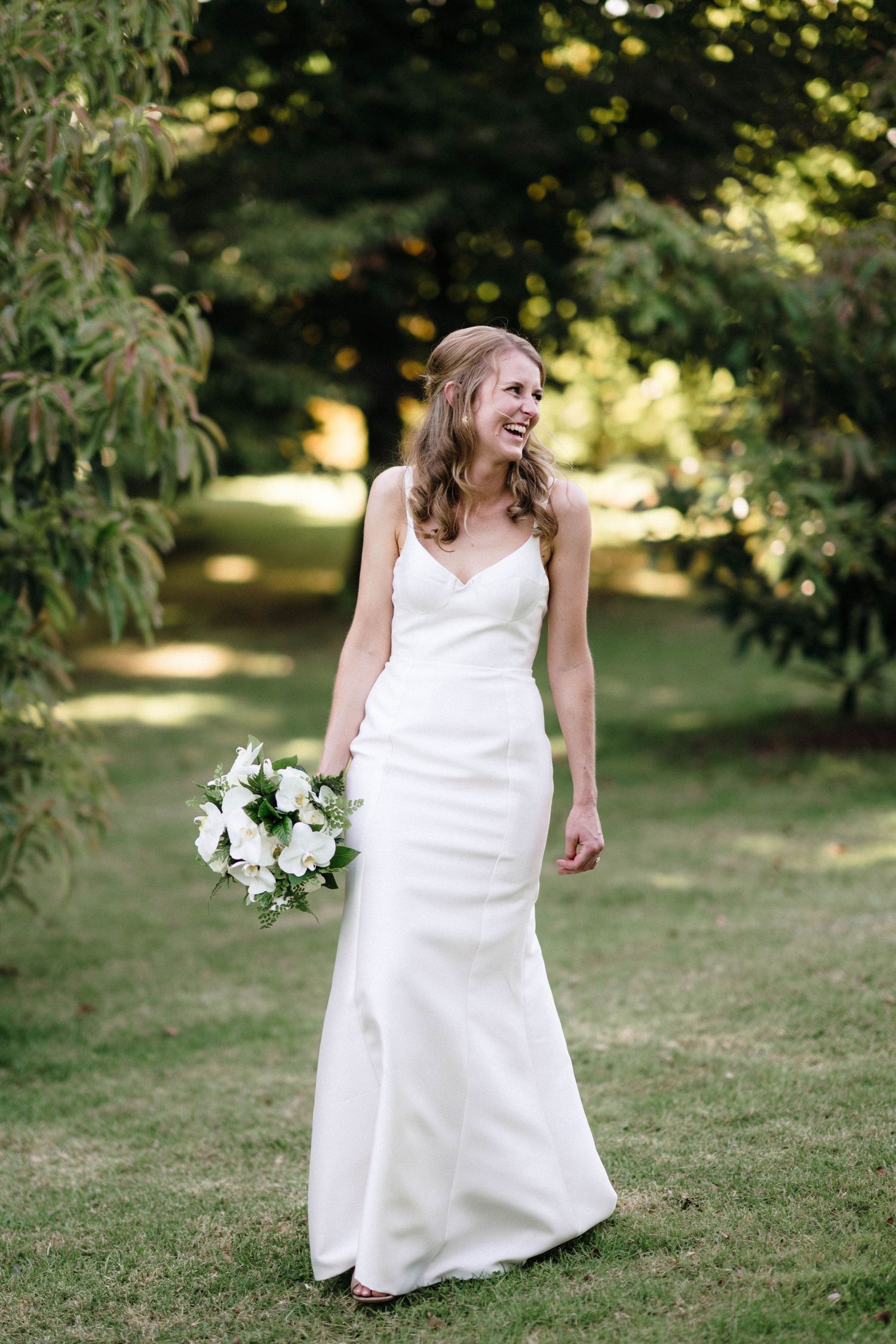 A bride in a white wedding dress poses with a bouquet in a sunlit garden setting.