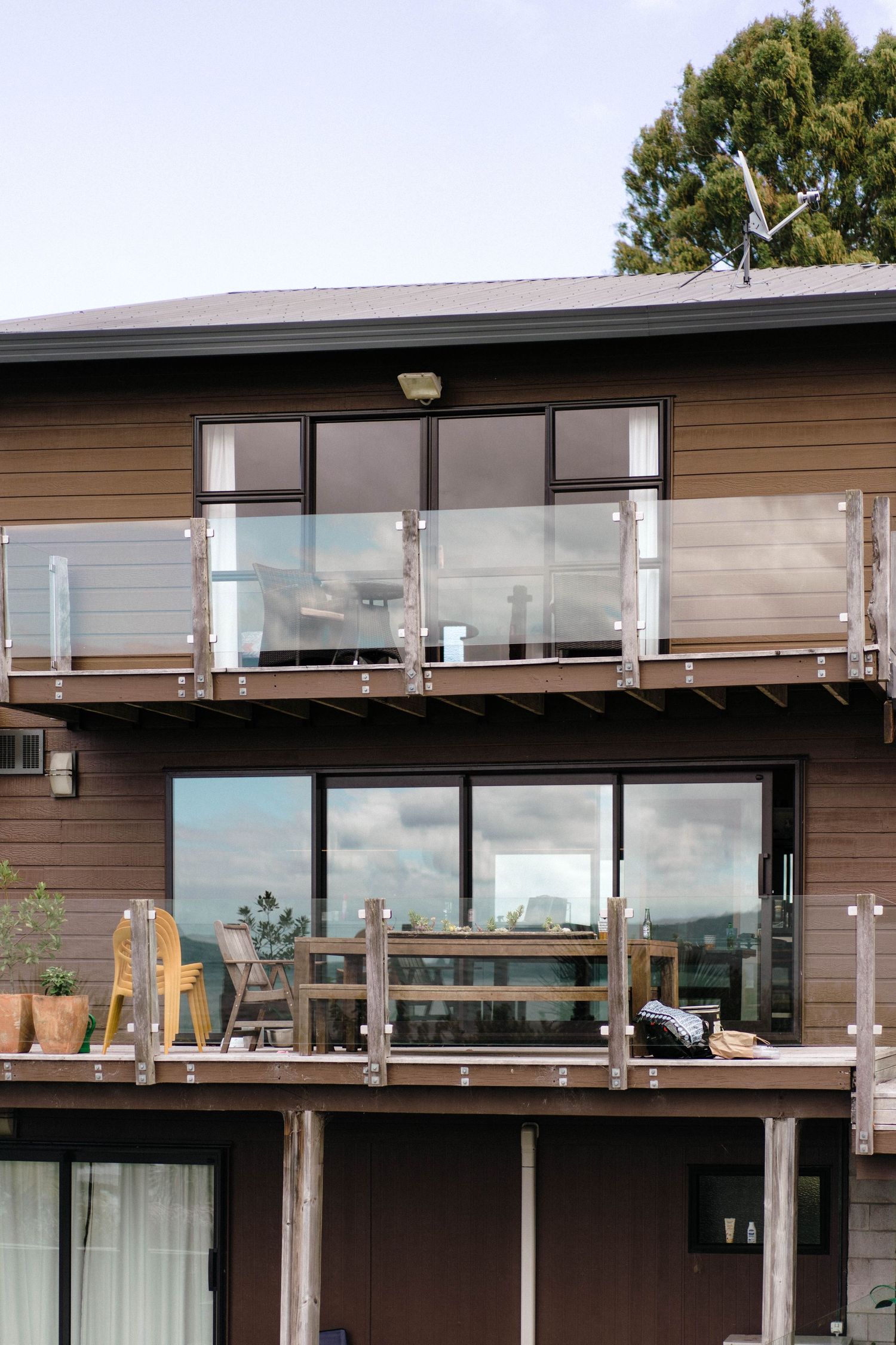 Modern two-story brown house exterior with glass balconies and wooden deck overlooking scenic view.