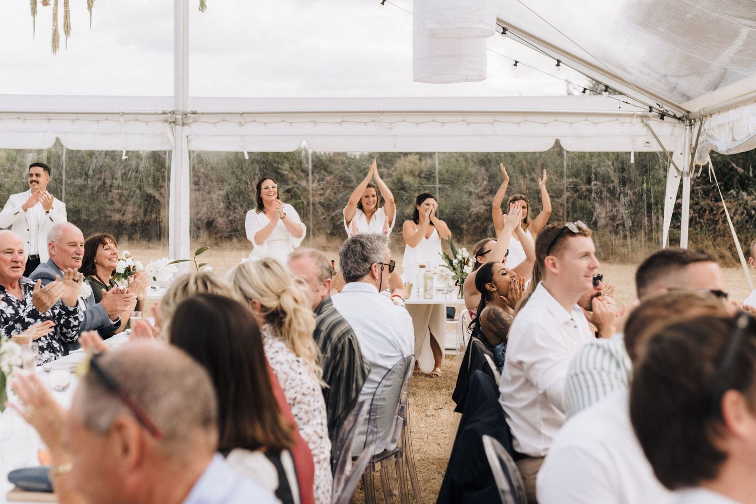 Wedding guests dancing and celebrating under a white tent at an outdoor reception.