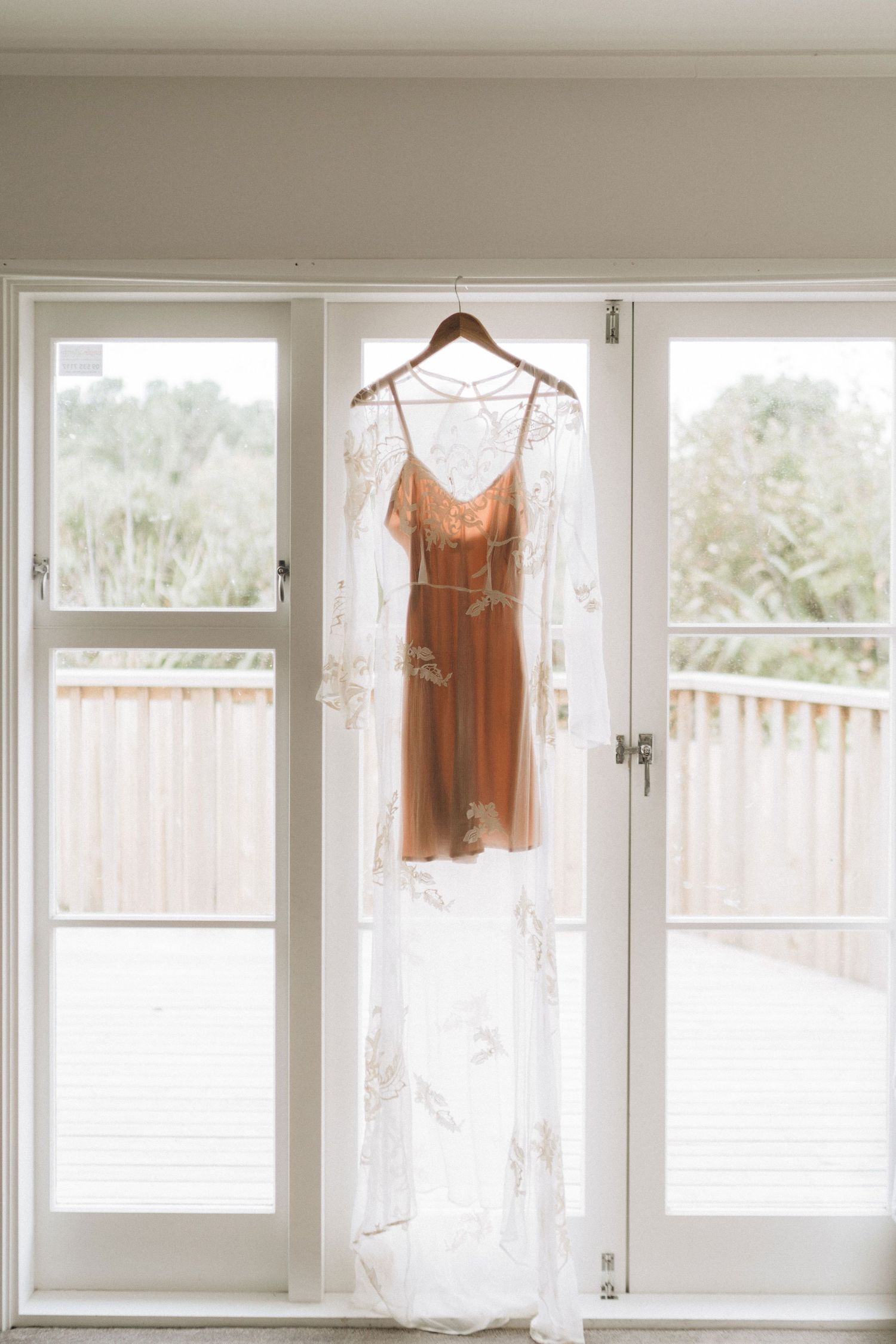 Multiple clothing items hanging in a bright window space against sheer white curtains.