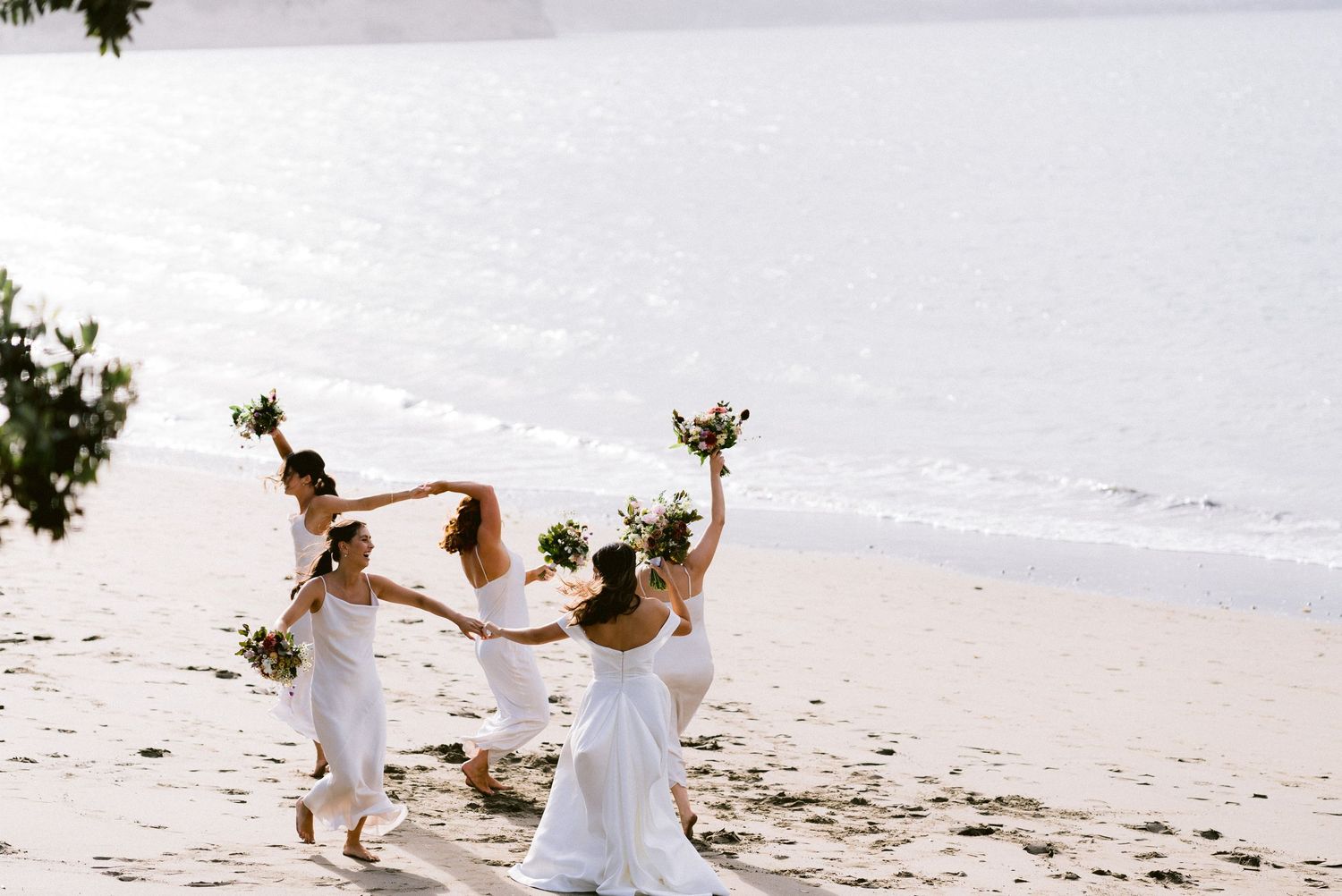 Wedding party members celebrating with raised bouquets on sunny beach.