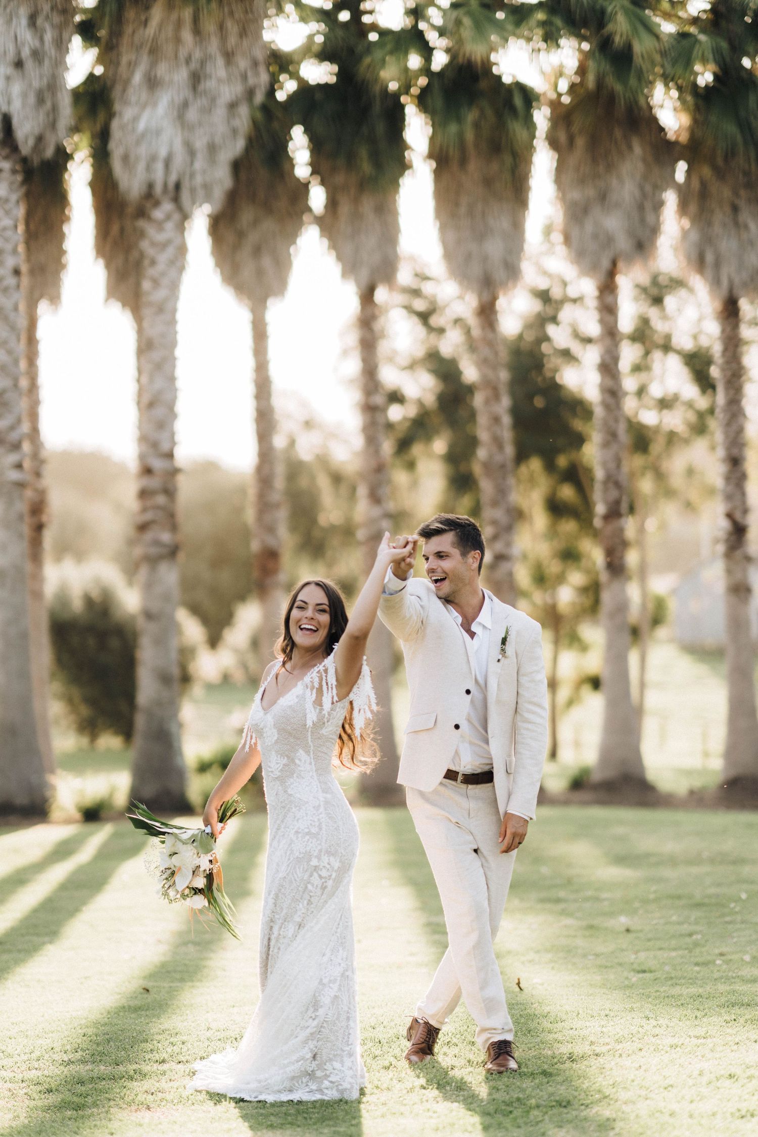 Newlyweds dancing and celebrating on grassy lawn beneath a row of majestic palm trees.