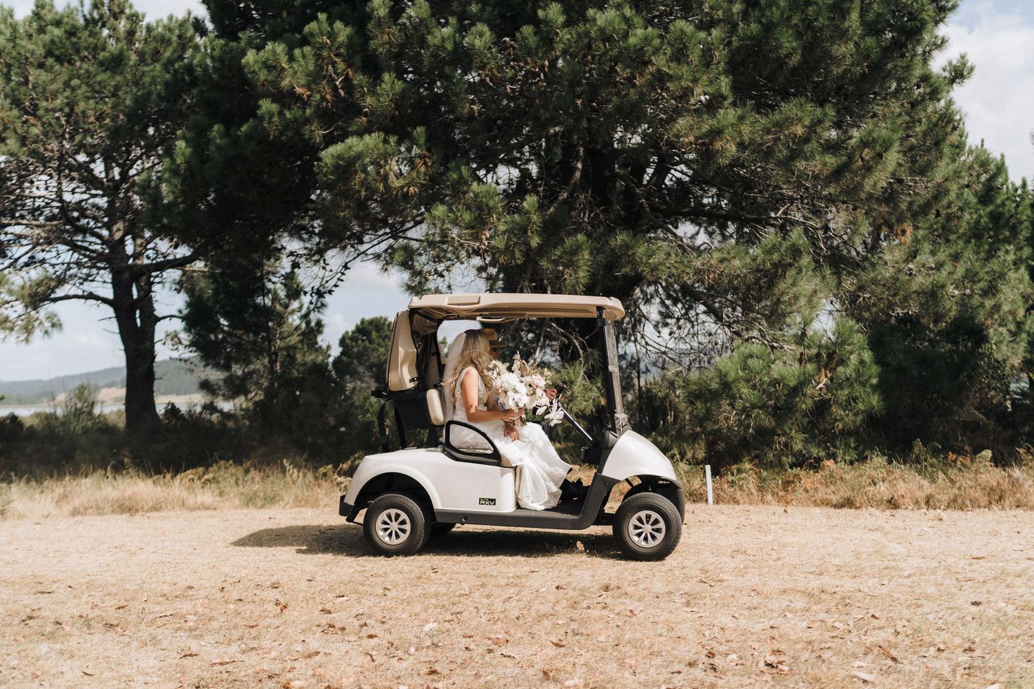 A white golf cart parked on a dirt path with evergreen trees in the background.