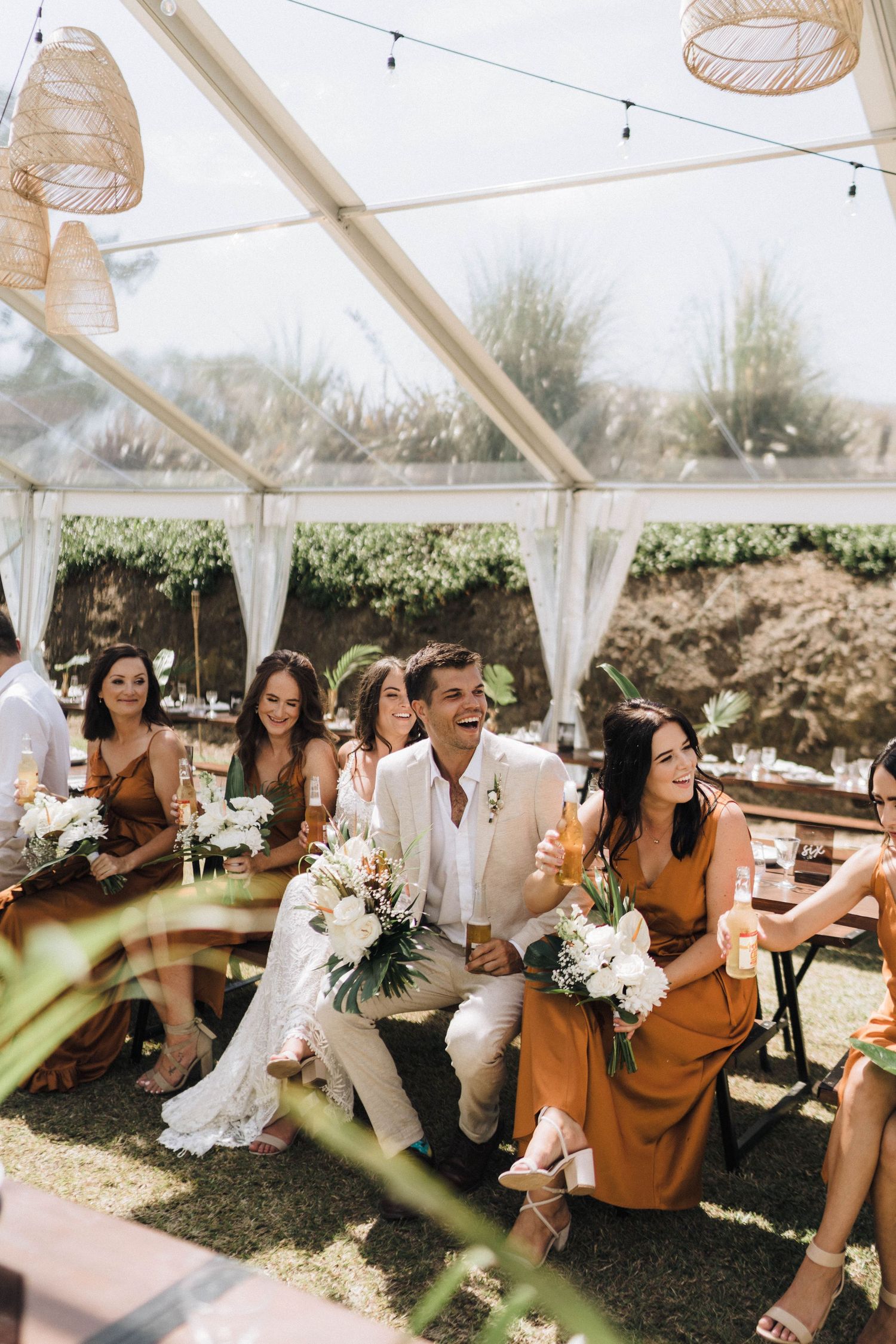 Wedding guests enjoy an outdoor celebration under a white tent canopy with hanging wicker lanterns and floral decorations.