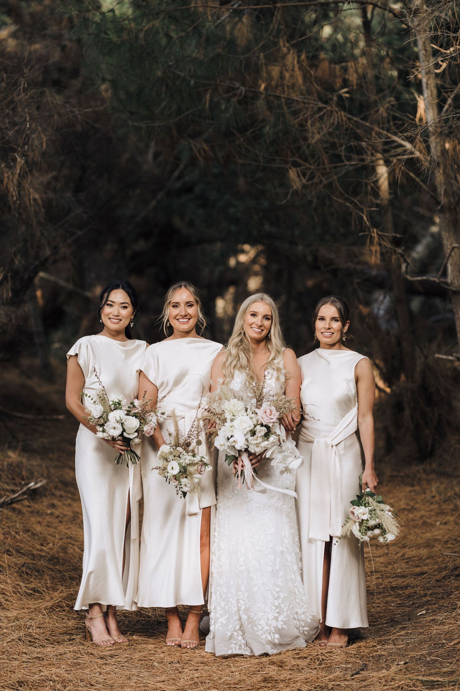 Bridesmaids in white dresses pose together holding elegant white floral bouquets in a forest setting.