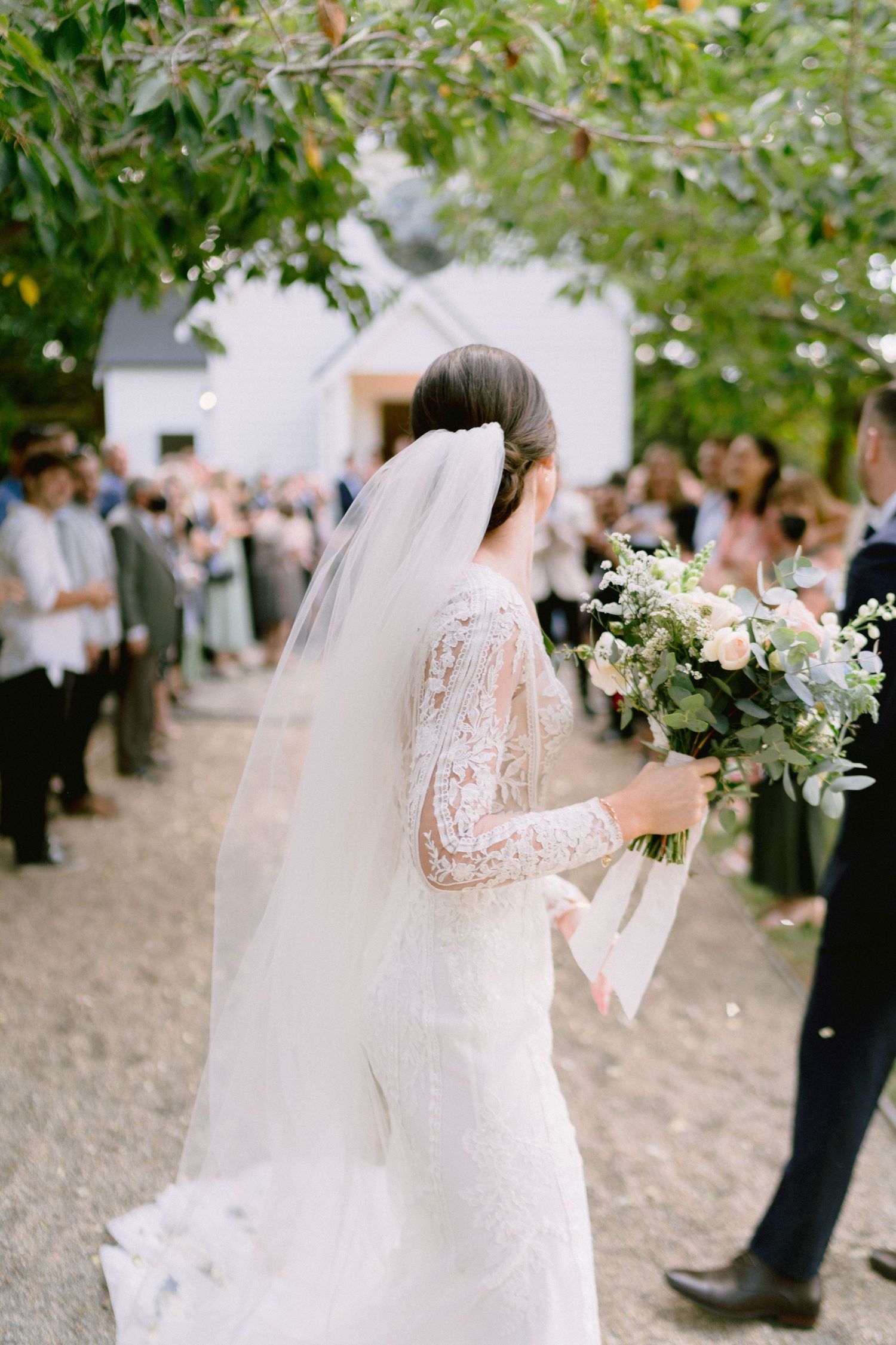 A bride in a lace dress walks down an outdoor path holding a white floral bouquet.