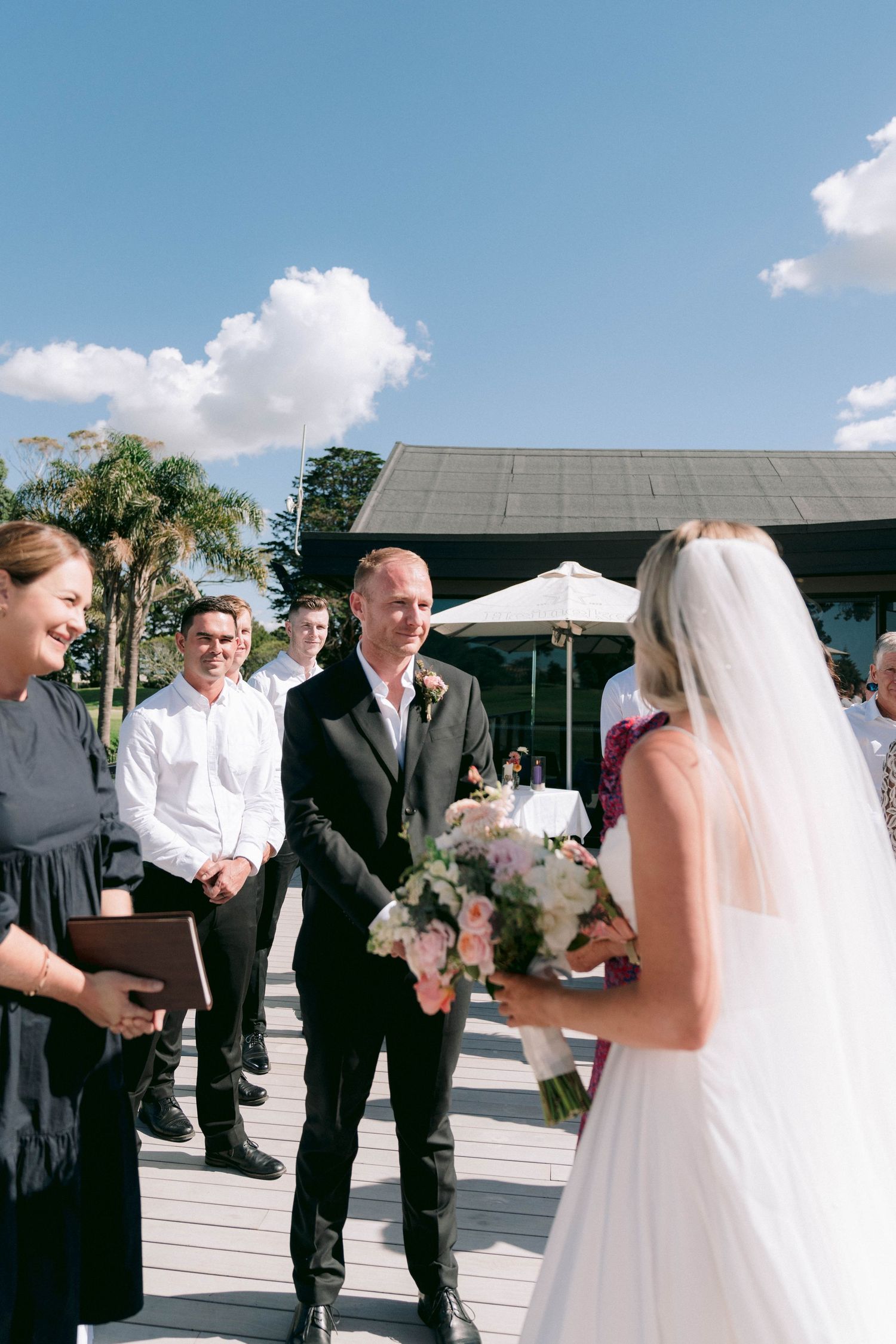 An outdoor wedding ceremony takes place on a deck under blue skies with white clouds.