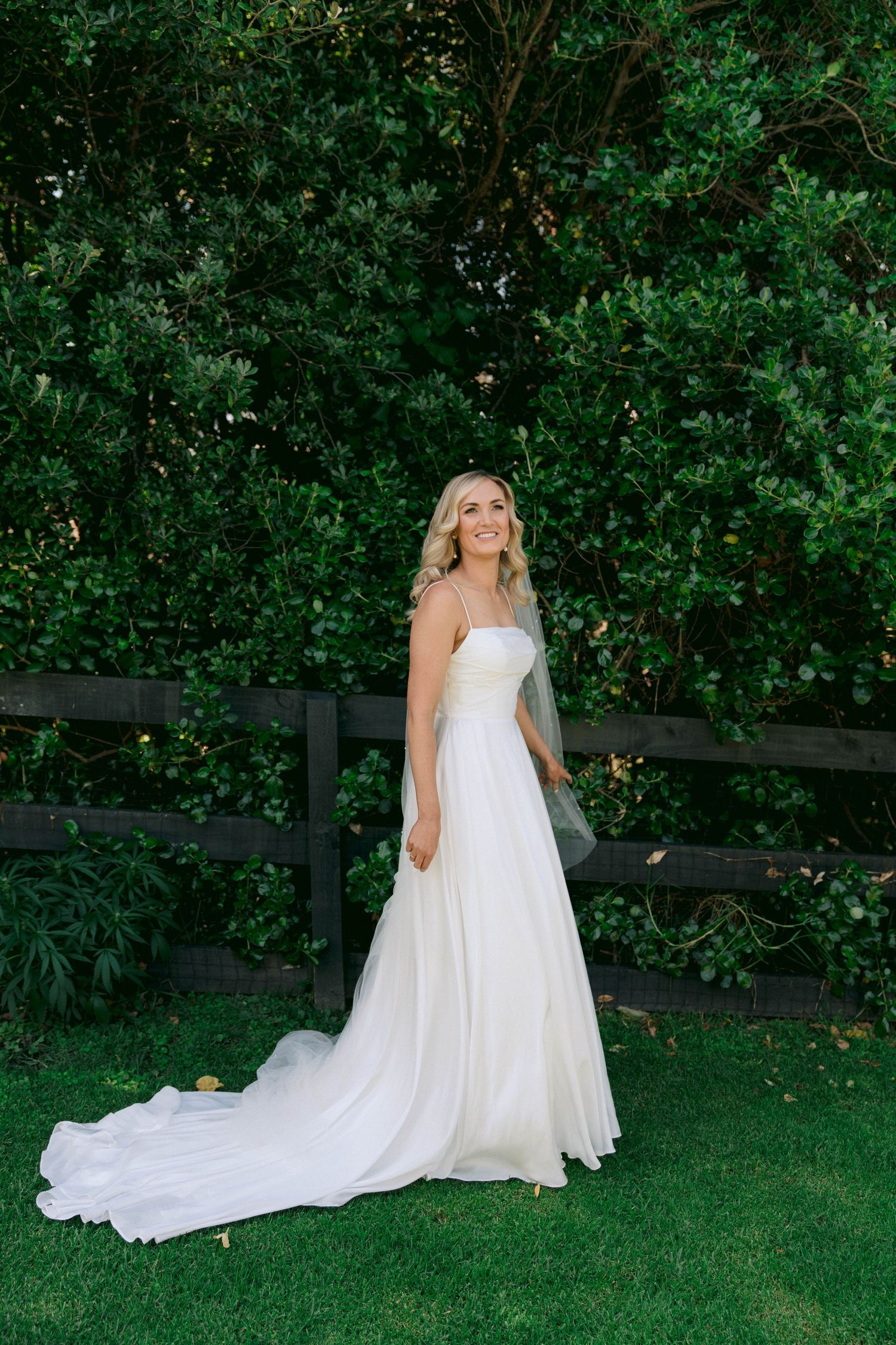 A stunning white wedding dress with a flowing train photographed against lush green foliage in an outdoor garden setting.