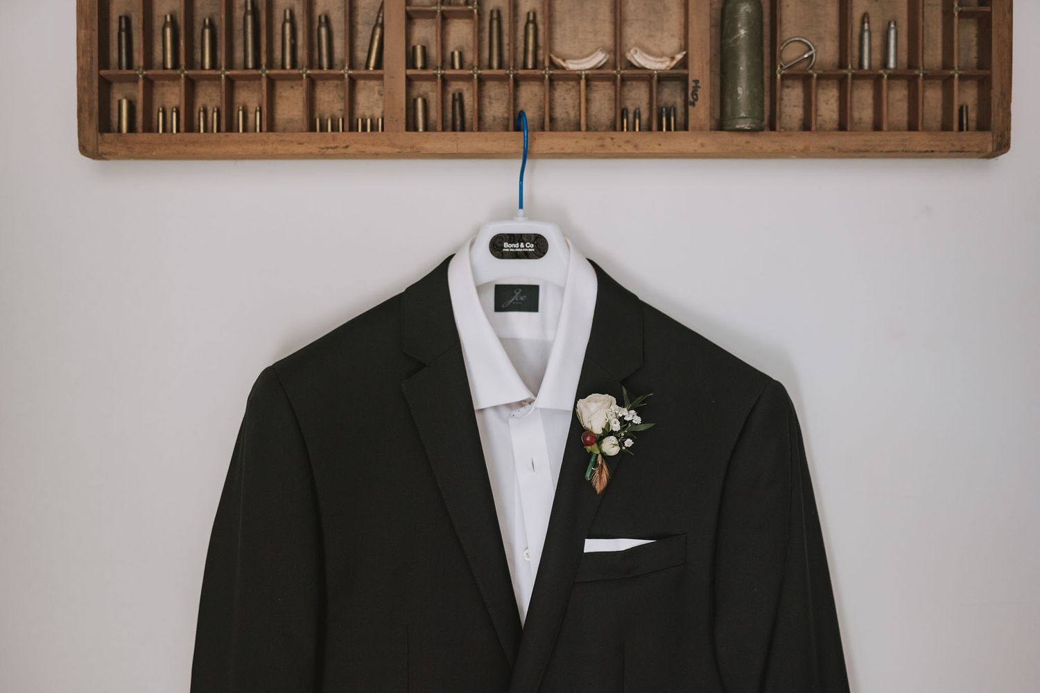 Black formal tuxedo jacket with white dress shirt and bridal boutonniere displayed on wooden hanger against light background.