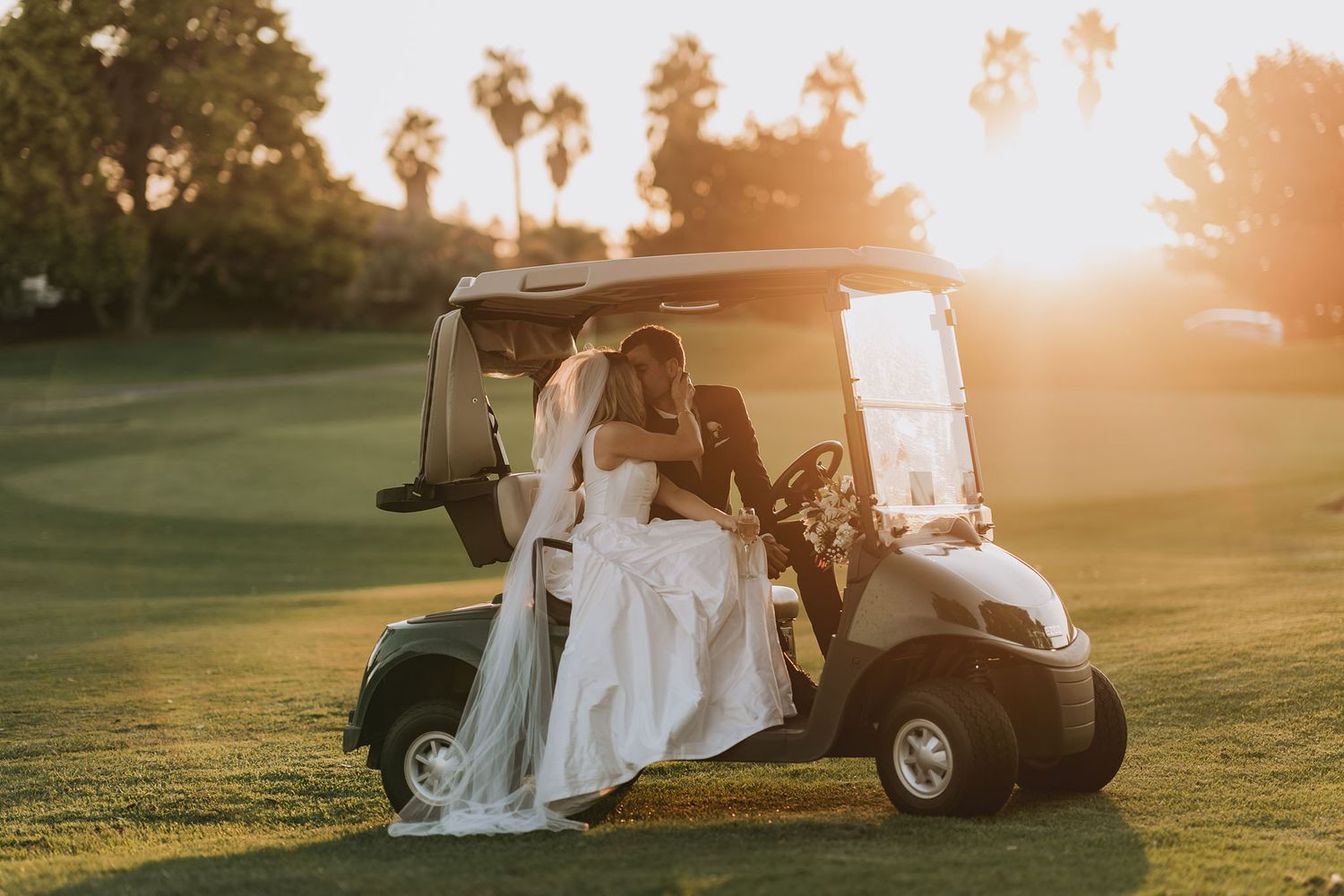 Newlyweds sitting together in golf cart at sunset on green golf course with palm trees in background.