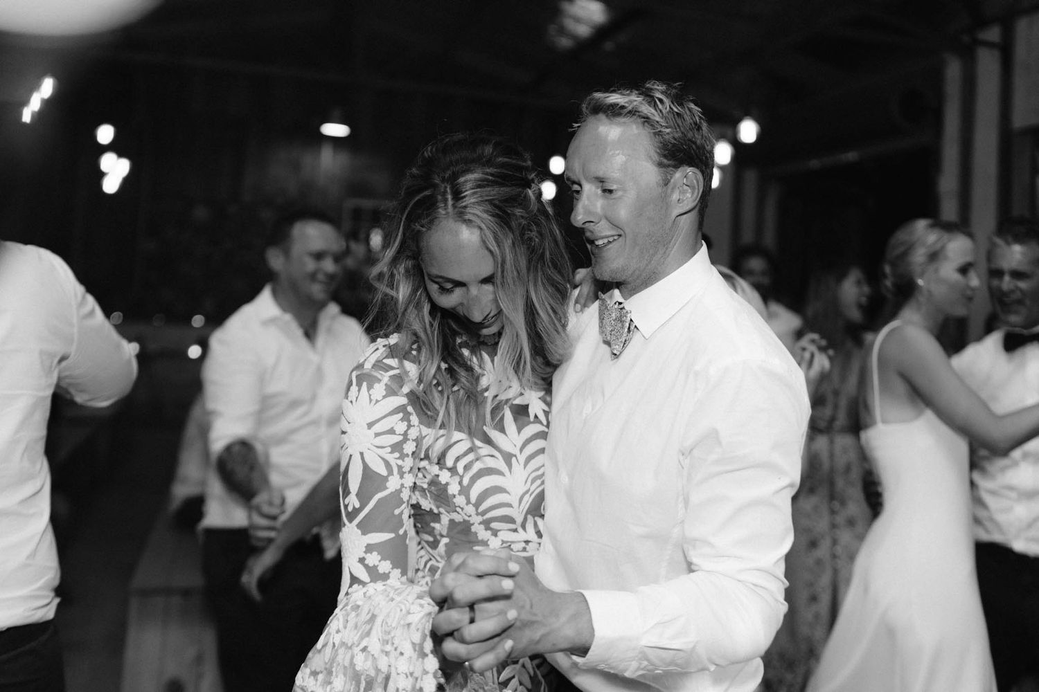 Black and white photo of a couple slow dancing together at a wedding reception with other guests visible in background.