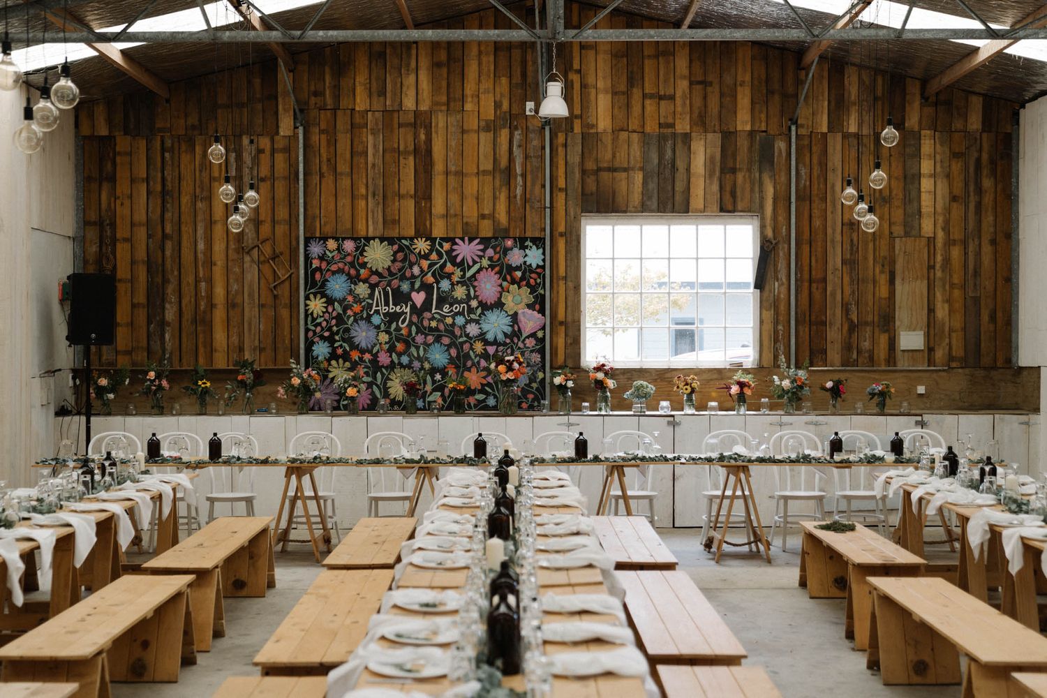 Wedding reception setup in a rustic barn with wooden tables and benches beneath string lights and floral decorations.