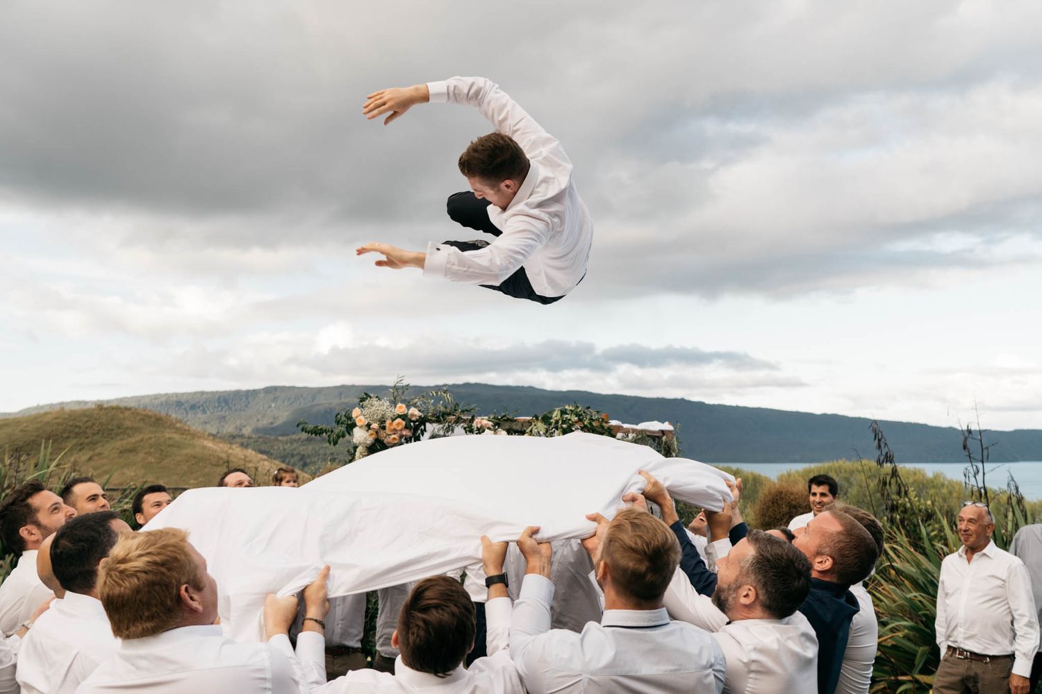 A group of people outdoors toss someone high into the air against a cloudy mountain landscape backdrop.