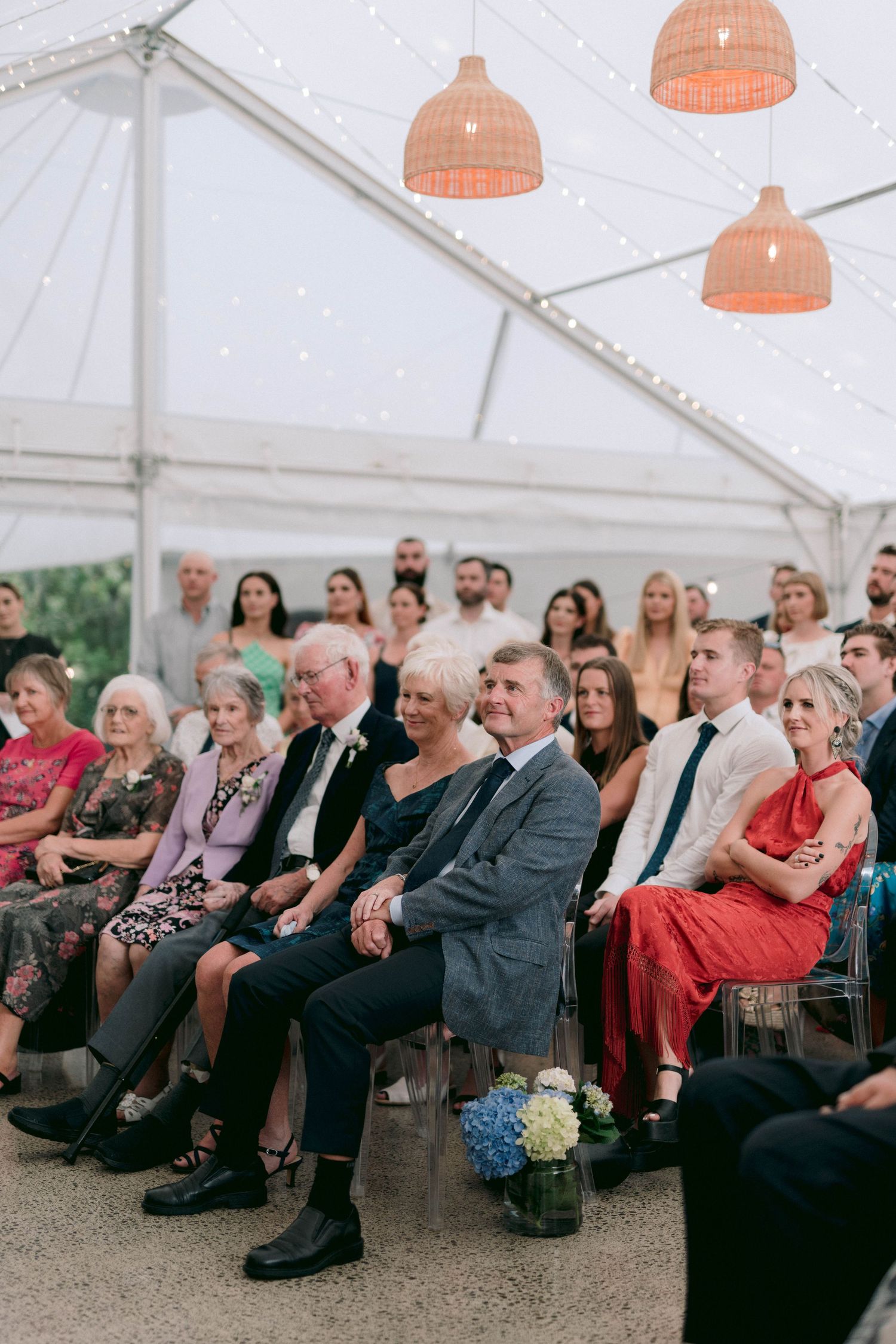 Wedding guests seated in rows under a white tent with coral pendant lights during outdoor ceremony.