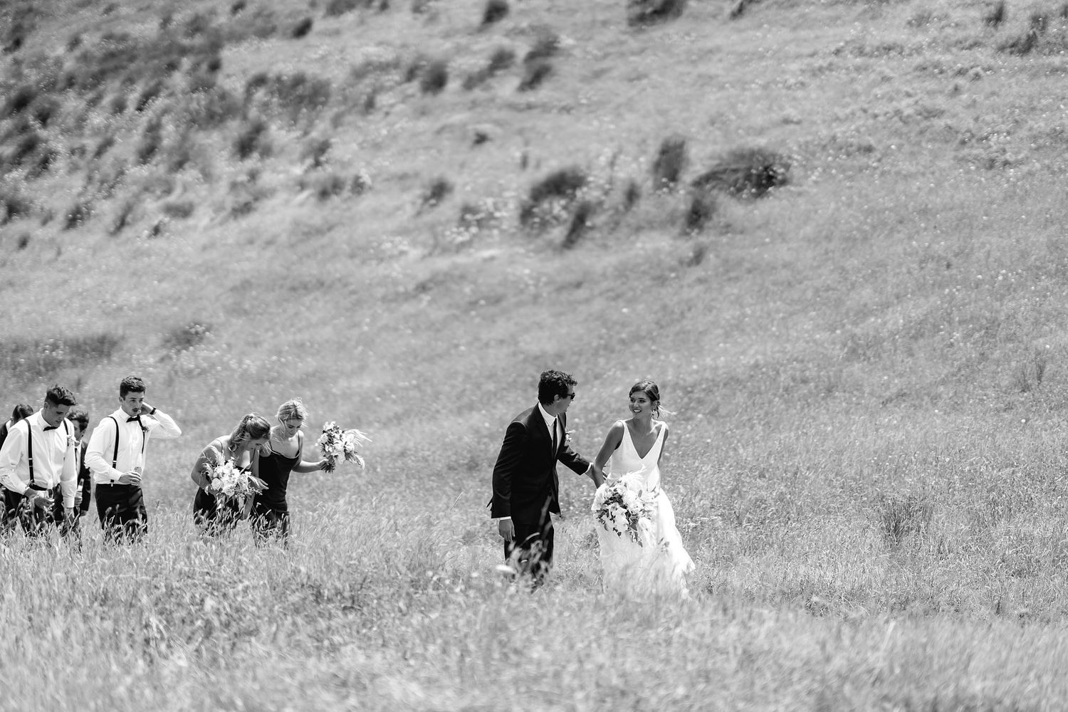 Black and white photo of a wedding couple walking through a grassy hillside with guests following behind.