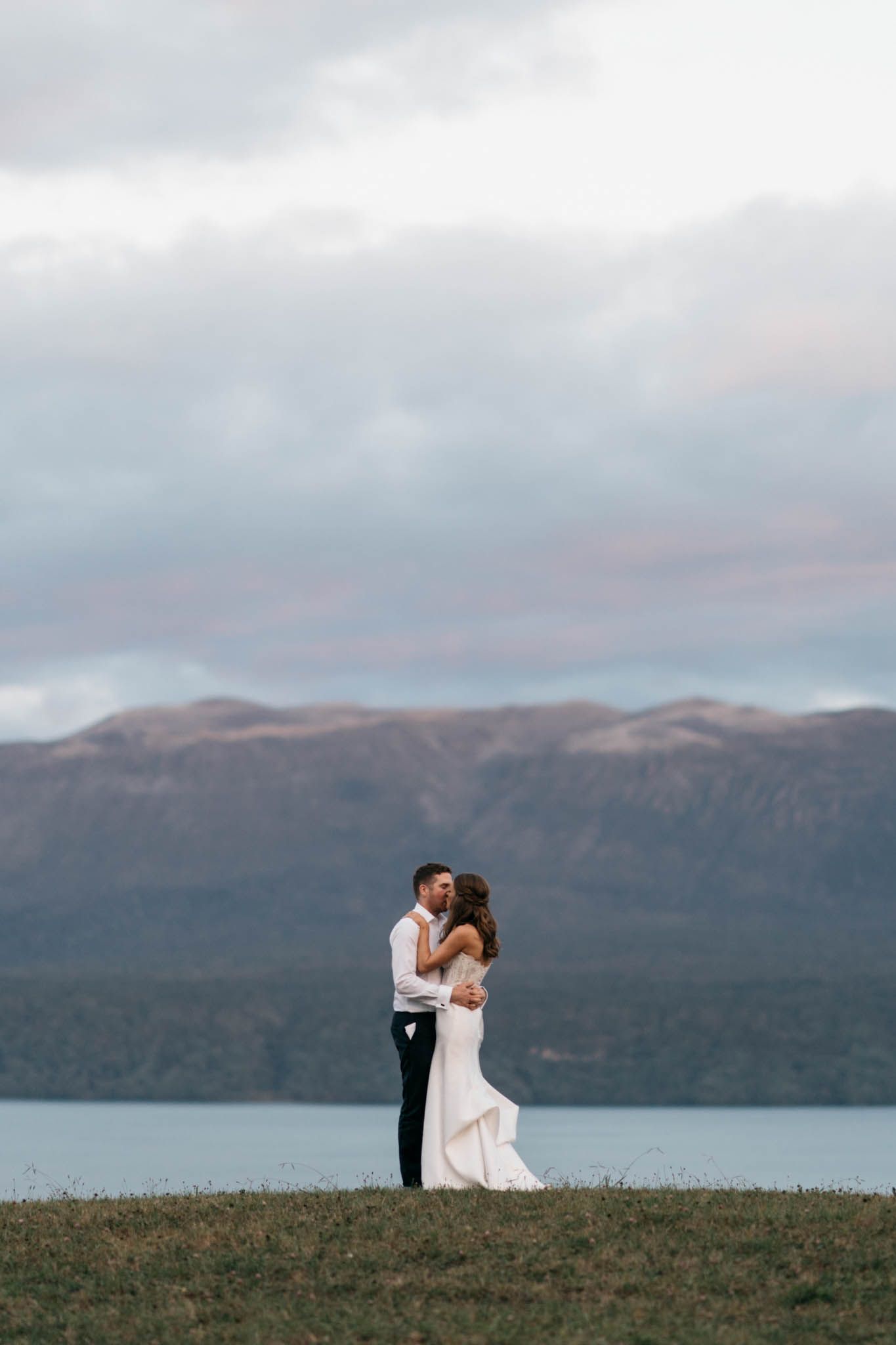 A couple stands silhouetted on a grassy hilltop against a dramatic mountain landscape beside a scenic loch in Scotland.