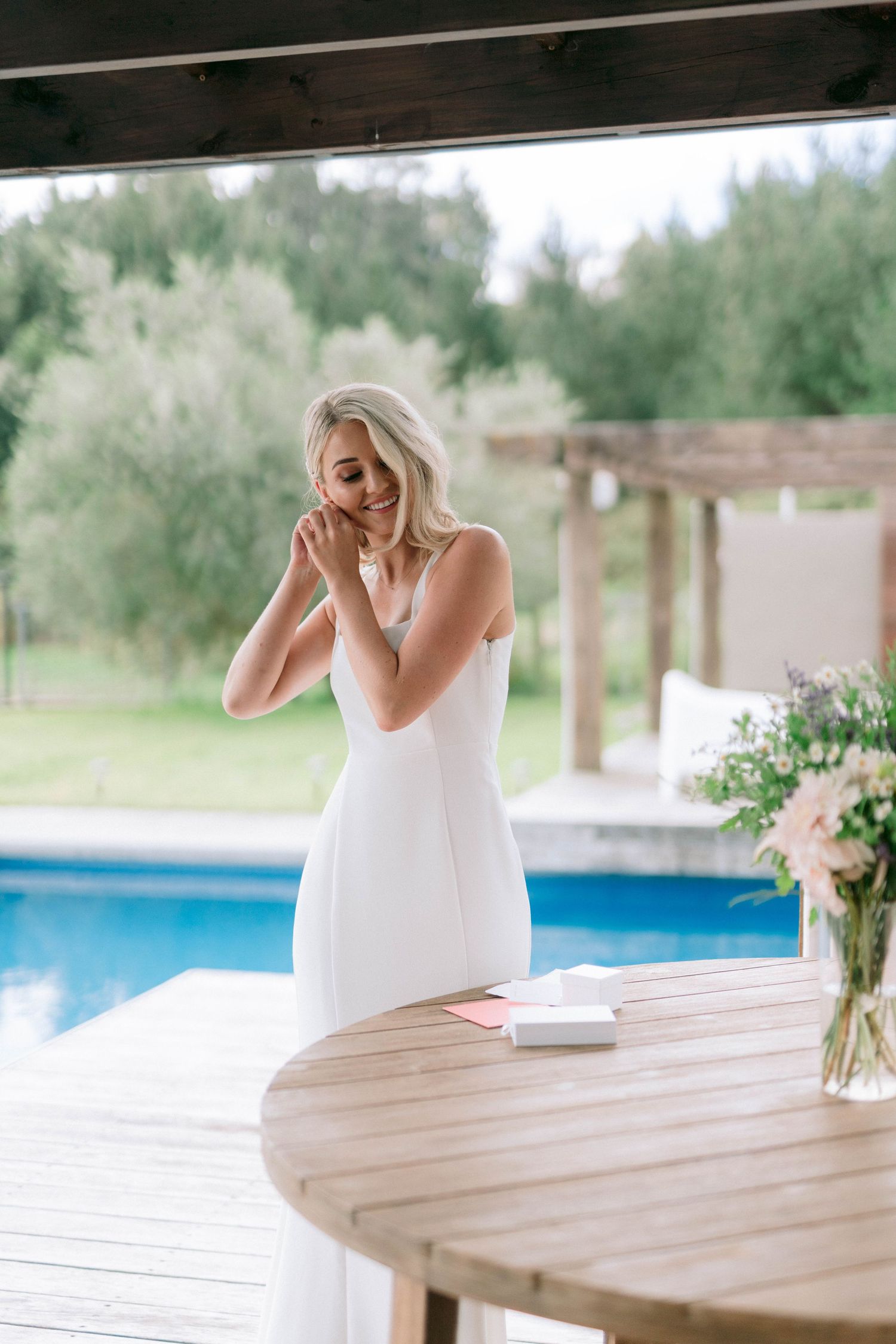 A woman in a white dress stands emotionally by a pool deck with a wooden table and floral arrangement.