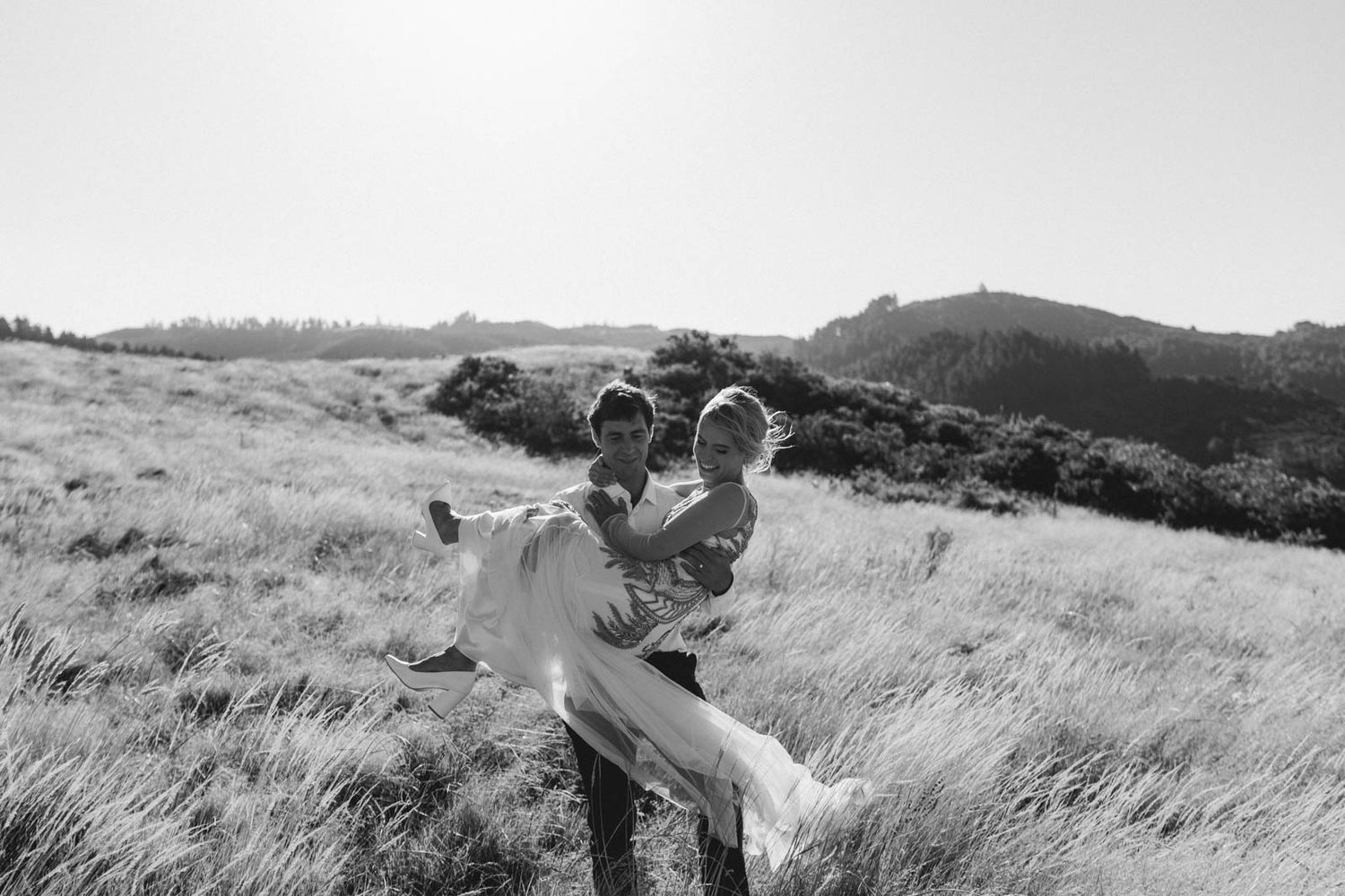 A loving grayscale moment captured in a scenic hilltop meadow with rolling hills in the background.