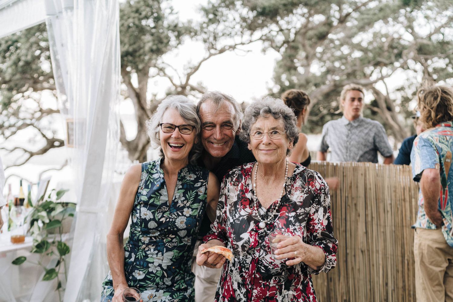 Three women smile together at an outdoor event with white curtains and bamboo decorations in the background.
