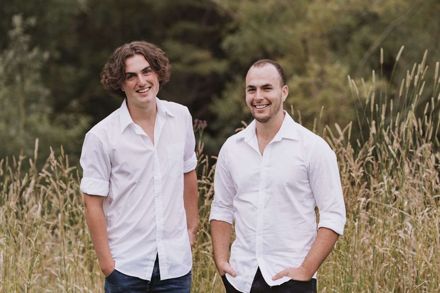 Two people wearing white button-down shirts stand together in a field of tall grass on a sunny day.