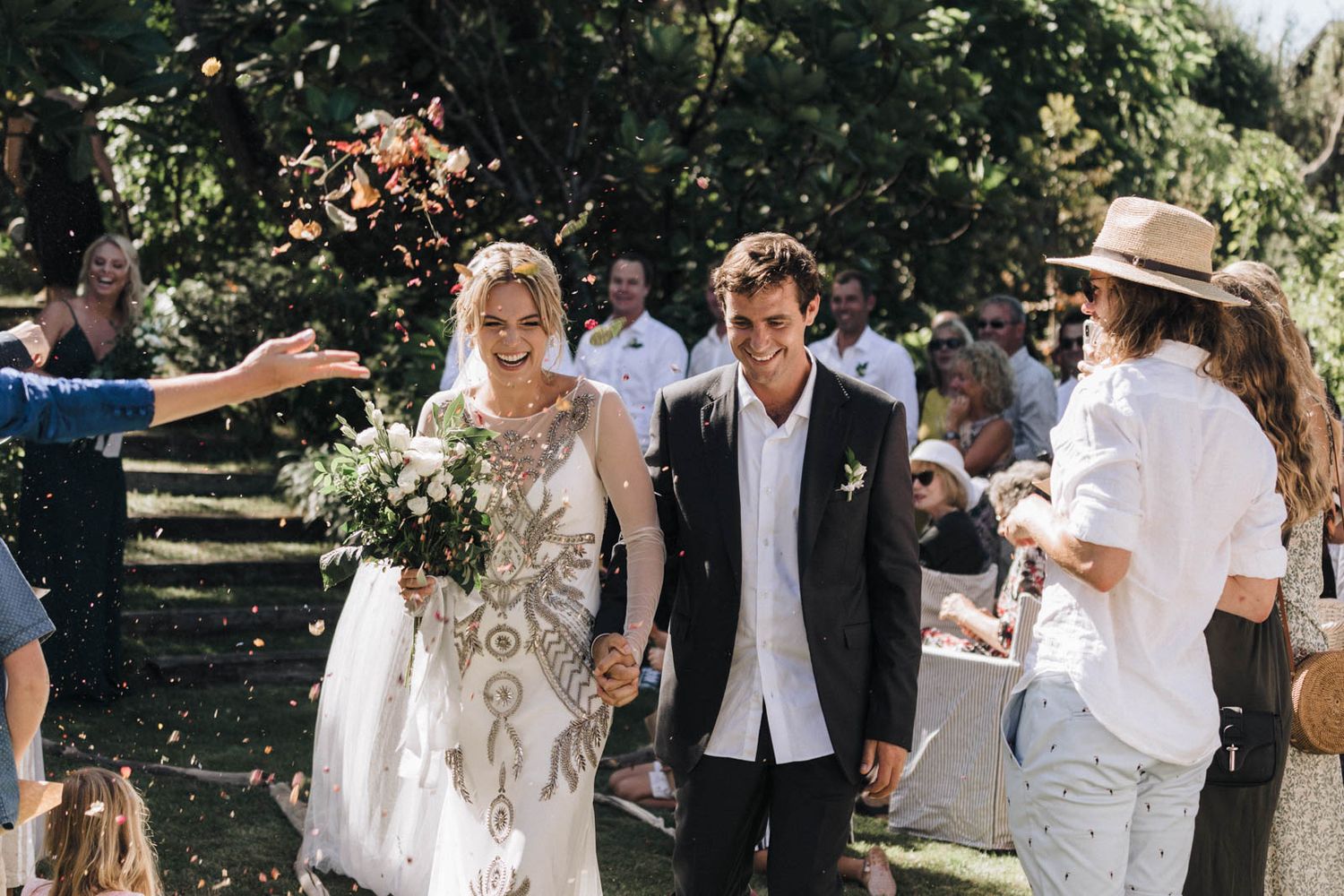 A couple walks down an outdoor aisle at a sunny garden wedding ceremony surrounded by guests and greenery.